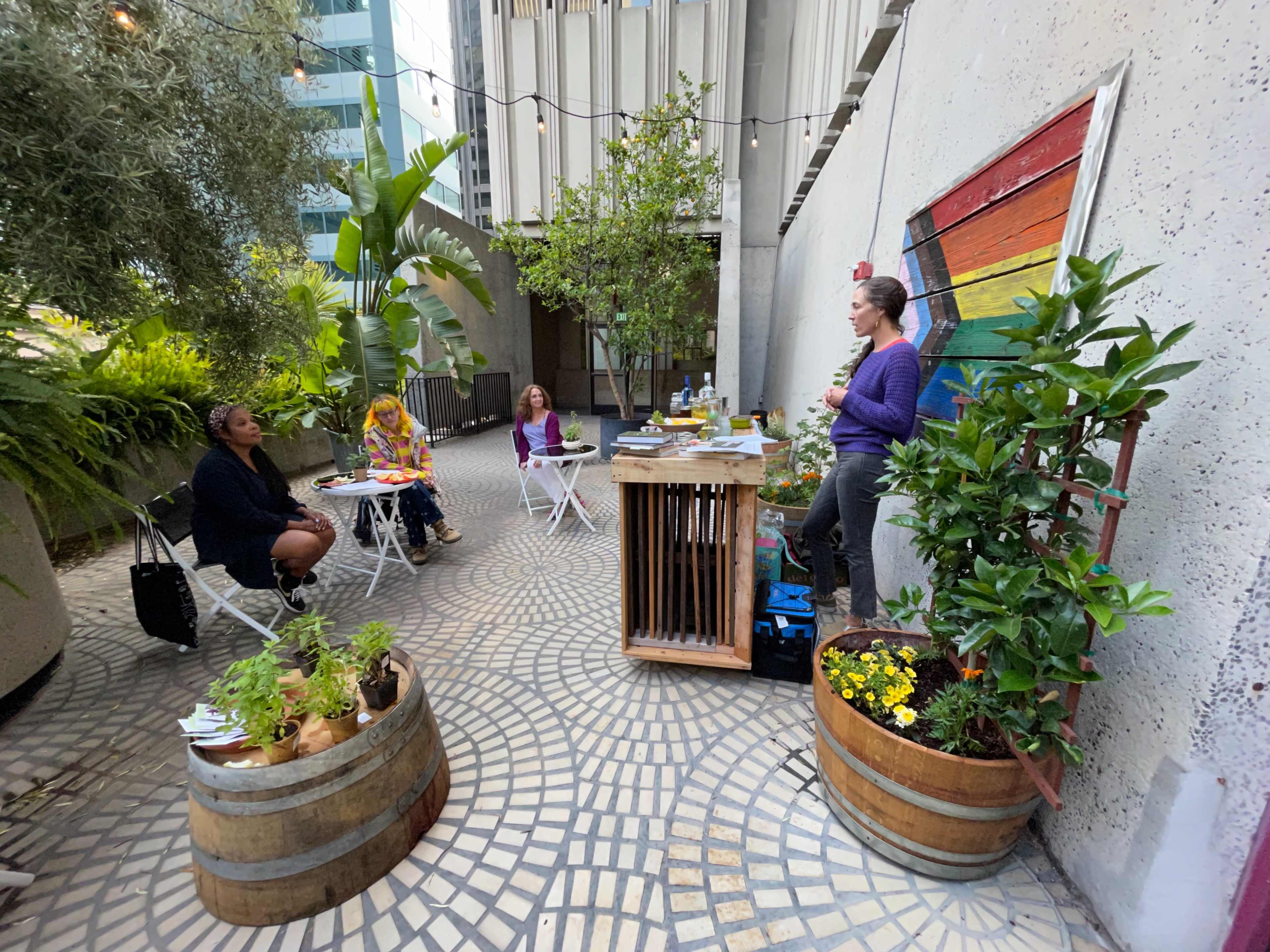 A woman stands by a decorated wall while a small group of people sits at white tables in a garden-like area filled with plants and containers.