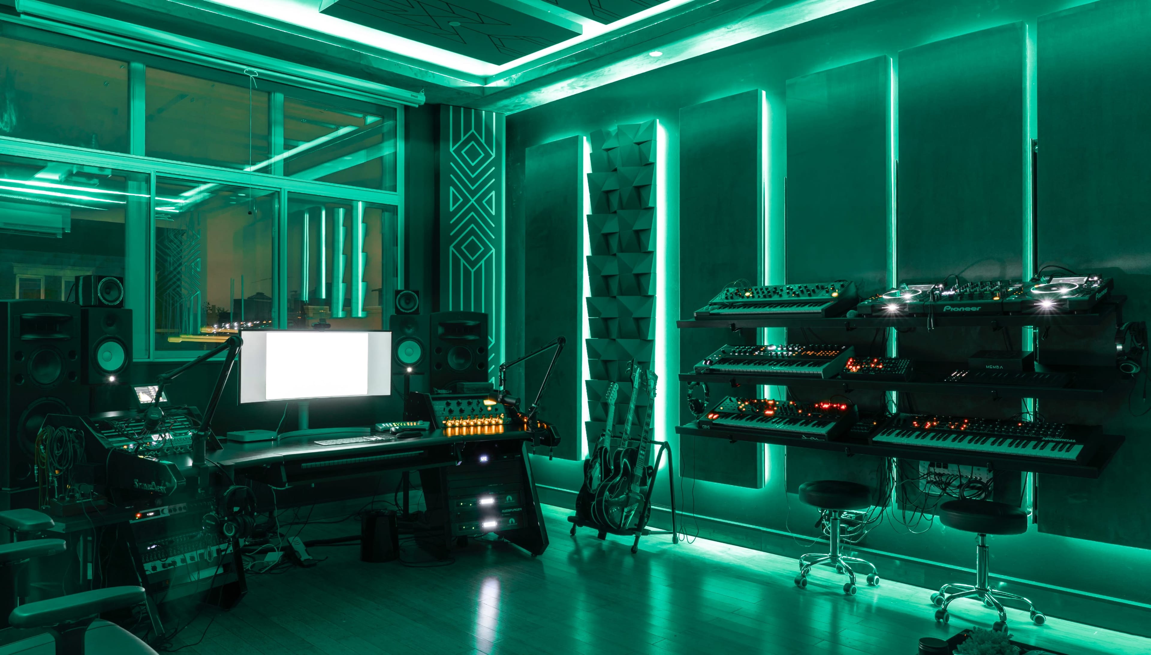 A music studio illuminated with green LED lighting, featuring a computer desk, various electronic instruments, and sound equipment along the walls.