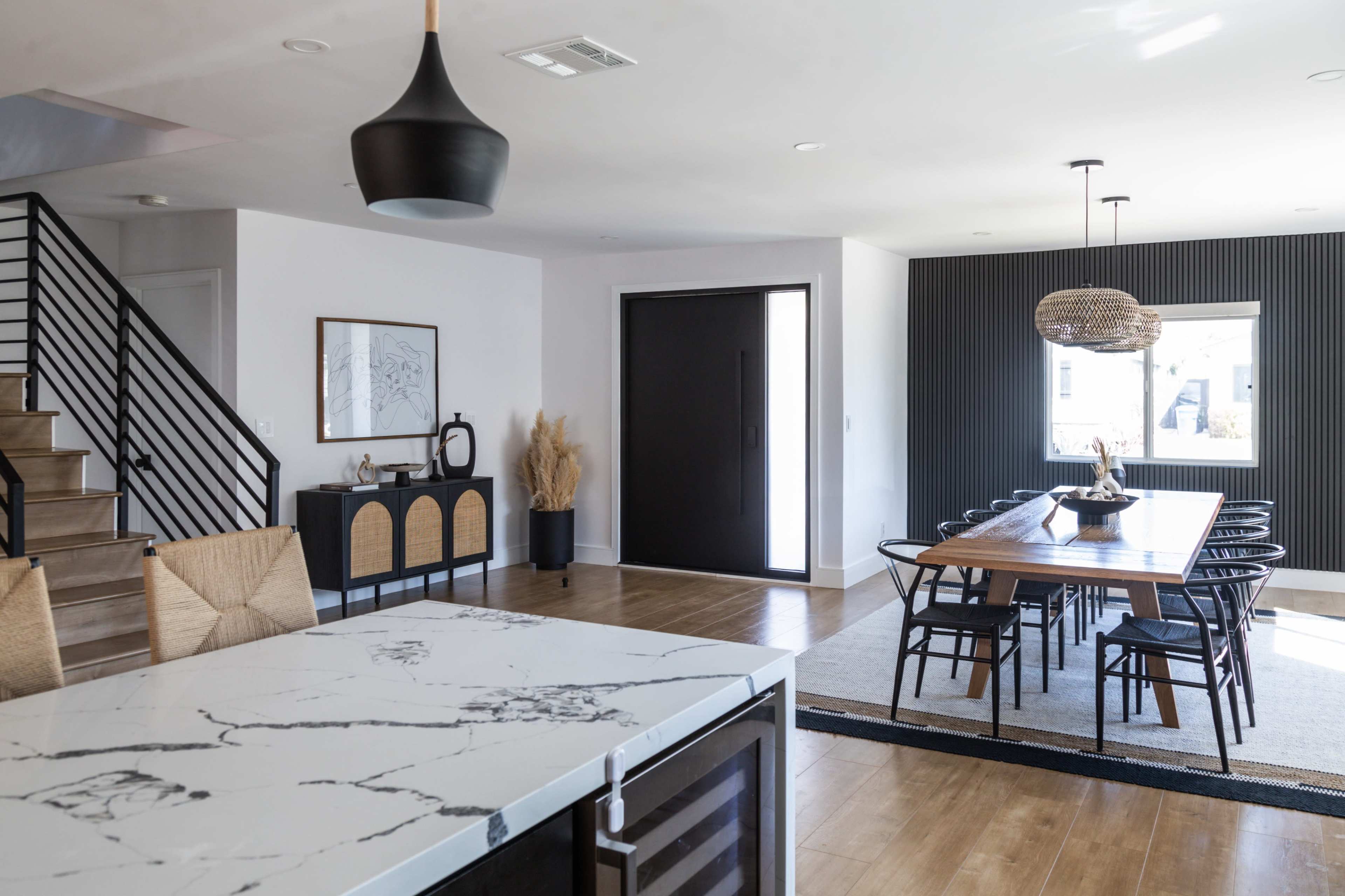 A modern interior features a dining area with a large wooden table, a kitchen island with a marble top, and a staircase leading to an upper level.