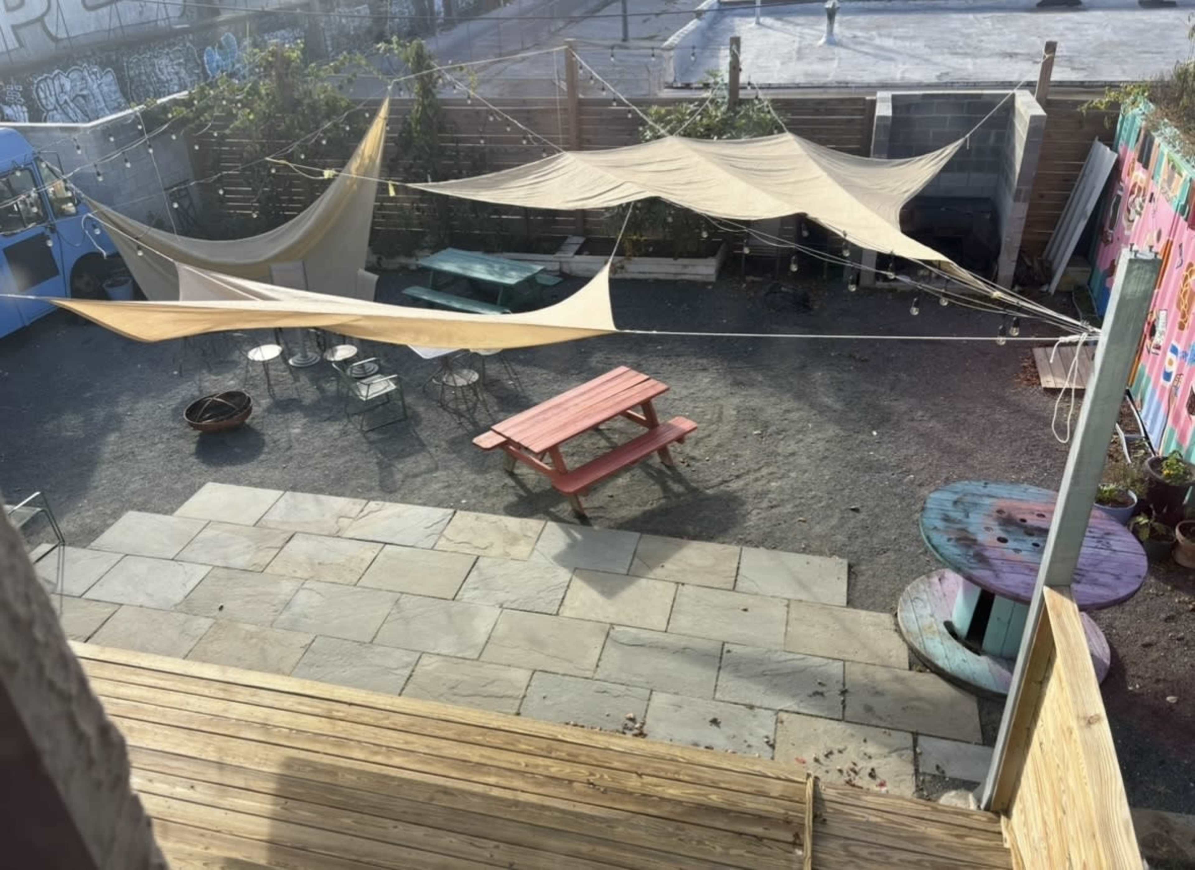 The image shows a backyard space featuring a wooden deck, a stone patio with a picnic table, and shade sails over a gravel area.