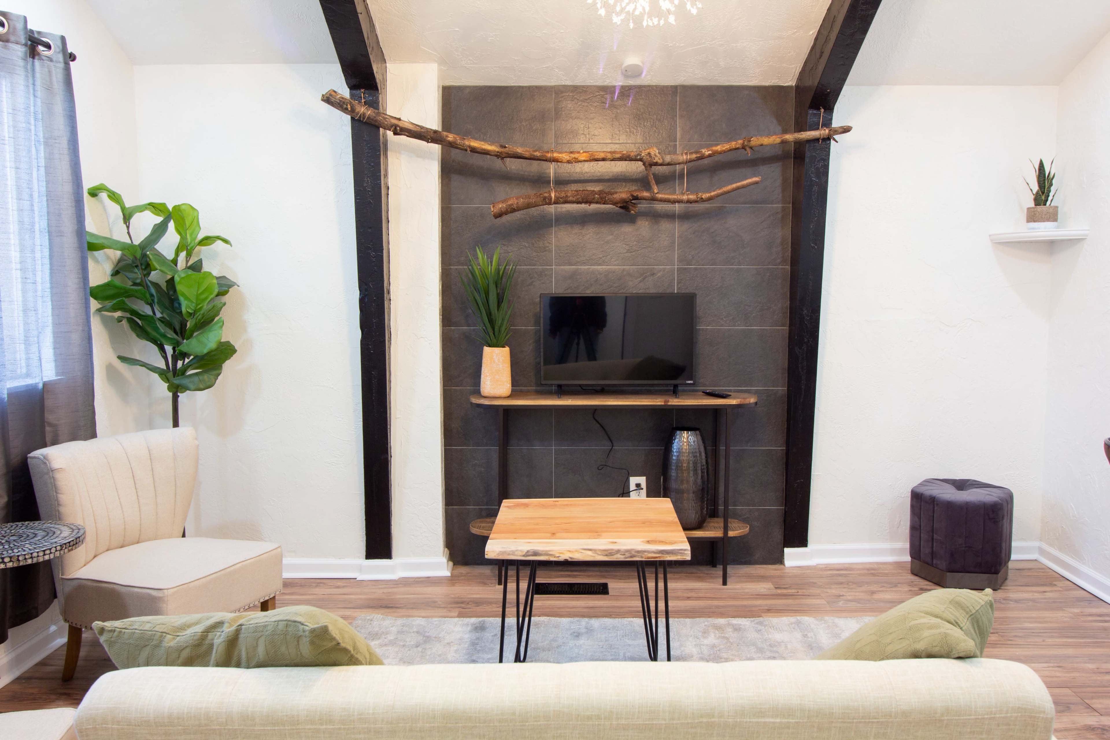 A living room featuring a green couch, a wooden coffee table, a television on a stand, and decorative plants against a light-colored wall with exposed beams.
