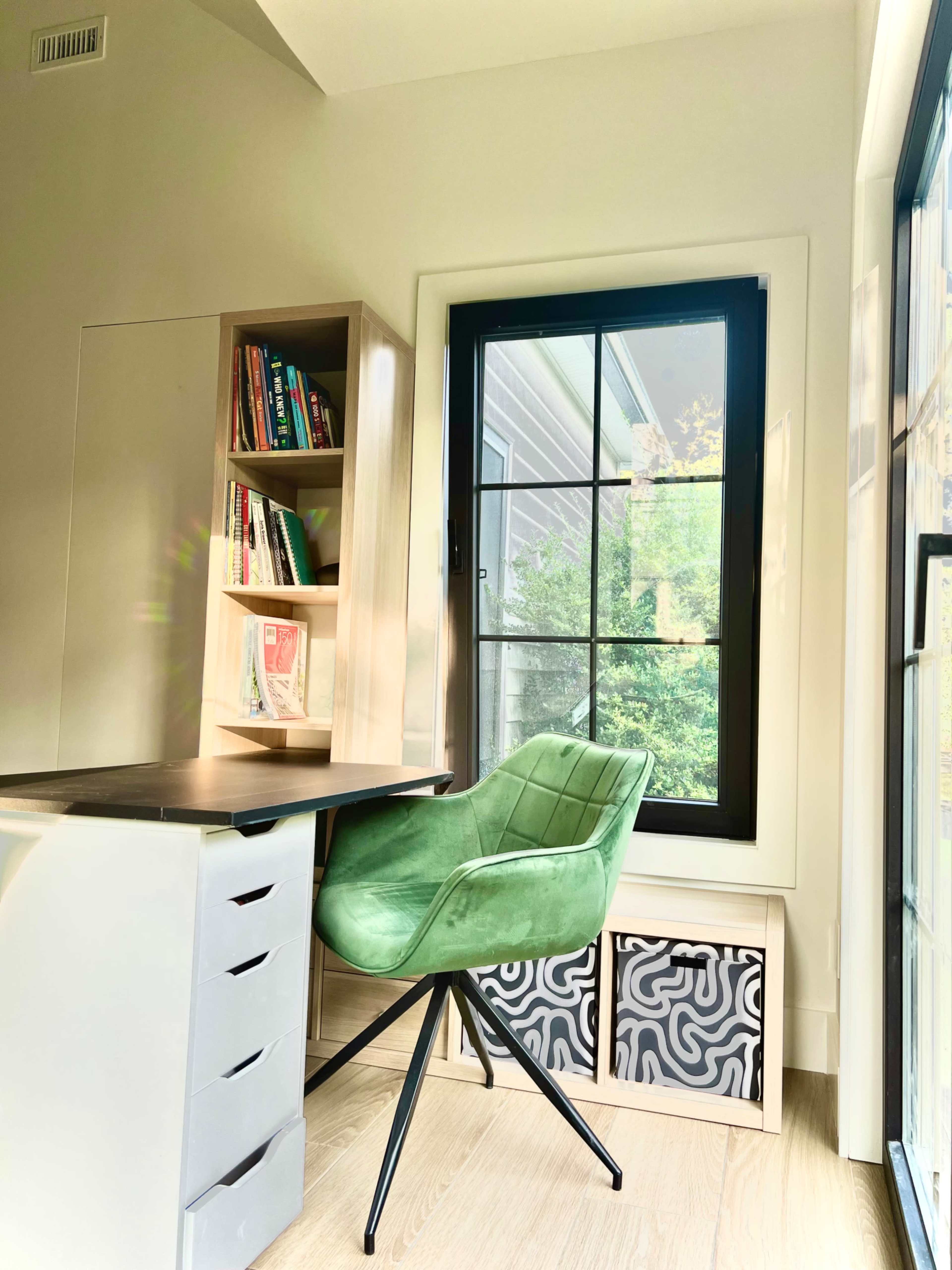 The image shows a modern workspace featuring a green chair, a desk with storage drawers, and a bookshelf filled with books, all positioned near a large window.