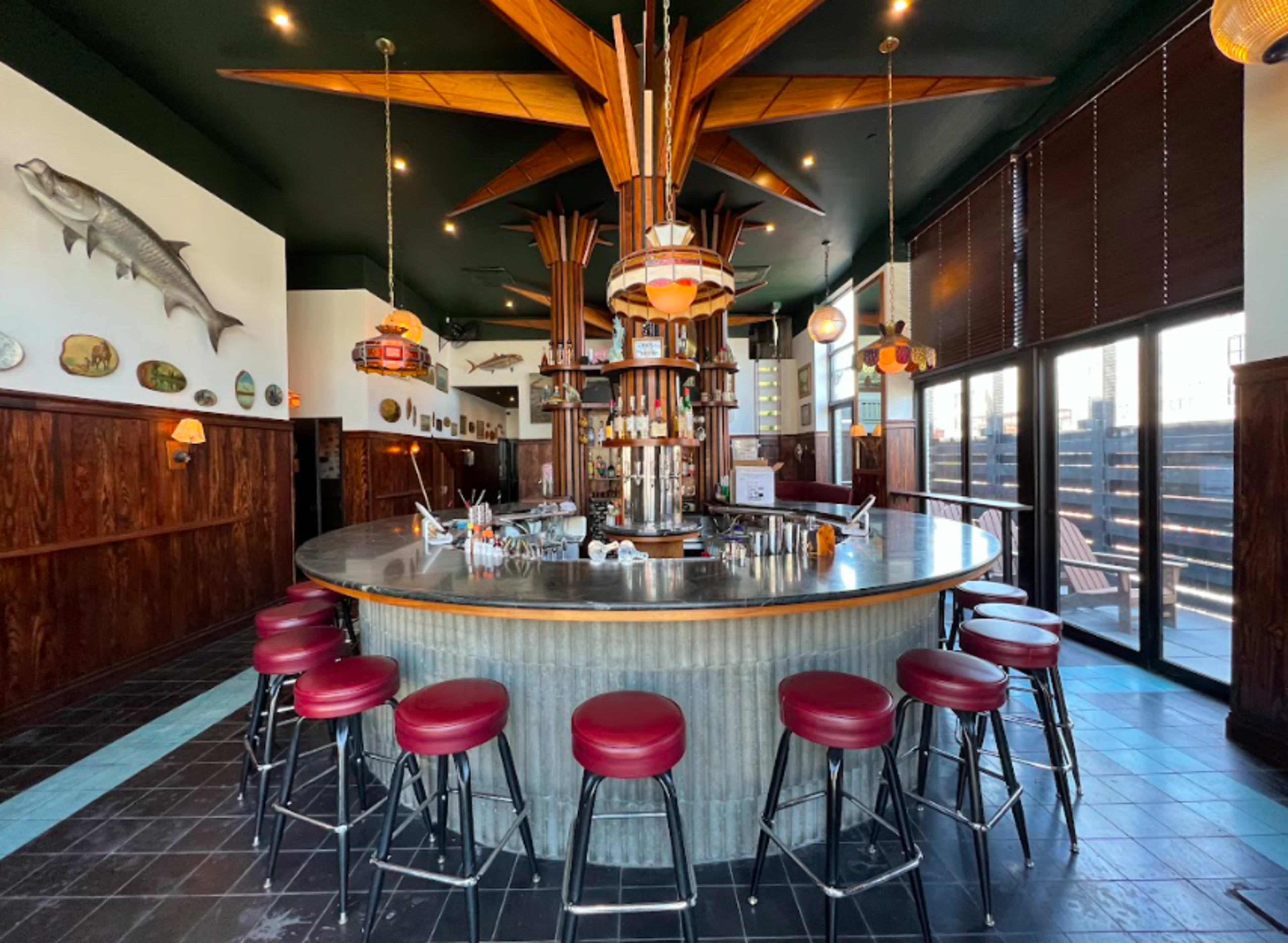 The image shows a circular bar with metal and wood elements, surrounded by red upholstered stools, in a restaurant featuring nautical decor and large windows.