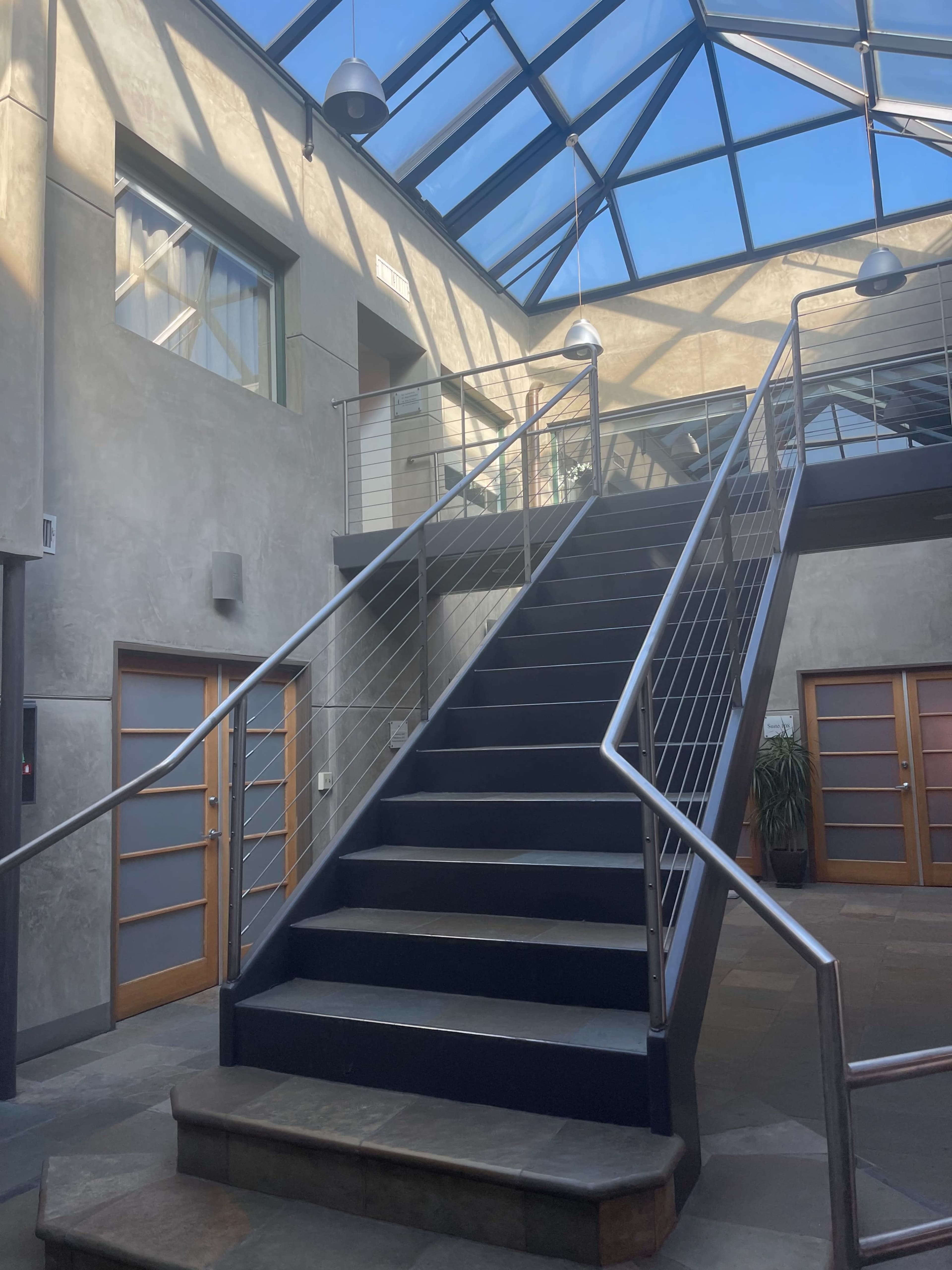 The image shows a staircase with a metal railing leading up to a second floor, surrounded by walls and a glass ceiling that allows natural light to enter the space.