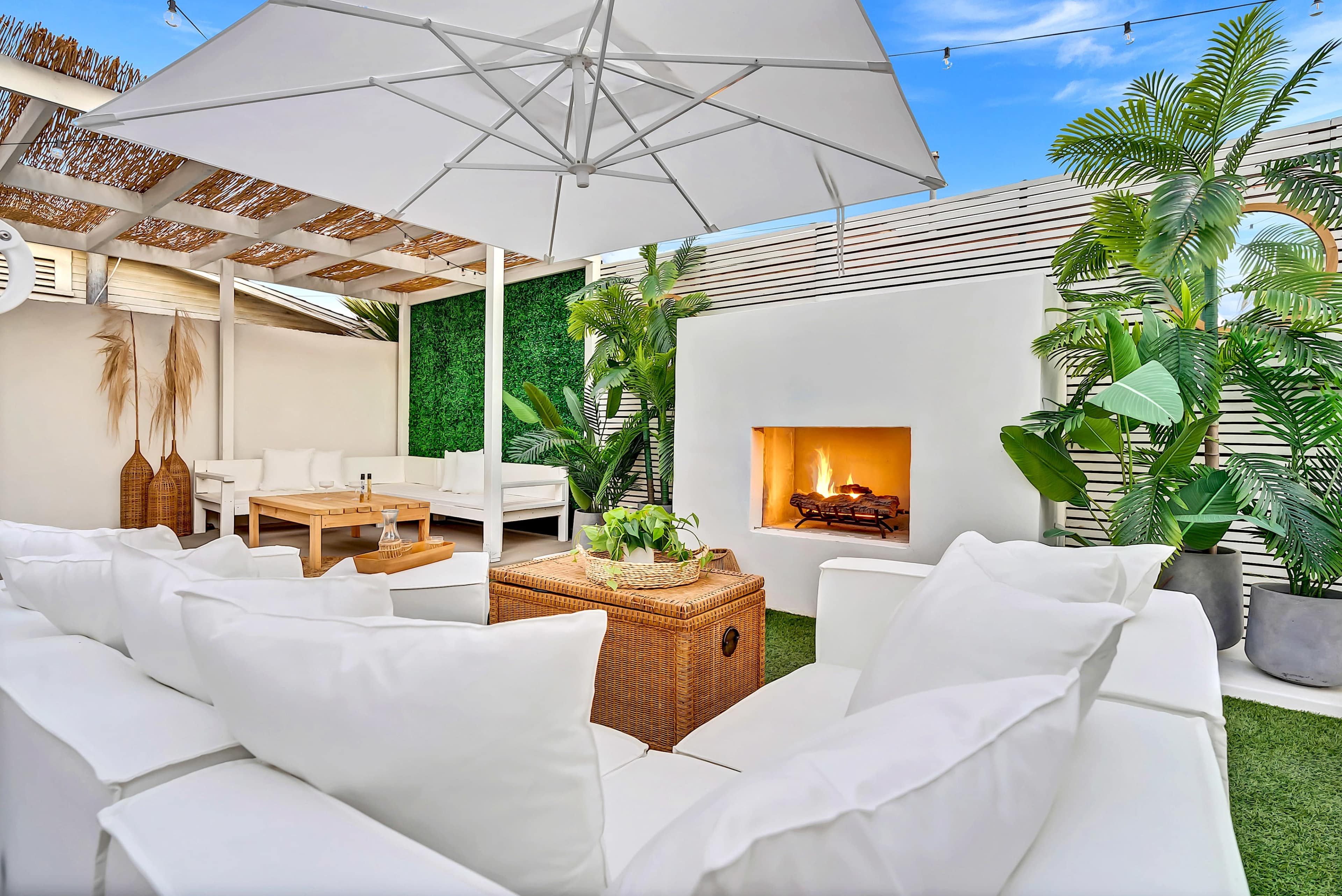 A cozy outdoor lounge area features a white sofa set, a wicker coffee table, an umbrella for shade, and a modern fireplace surrounded by lush greenery.