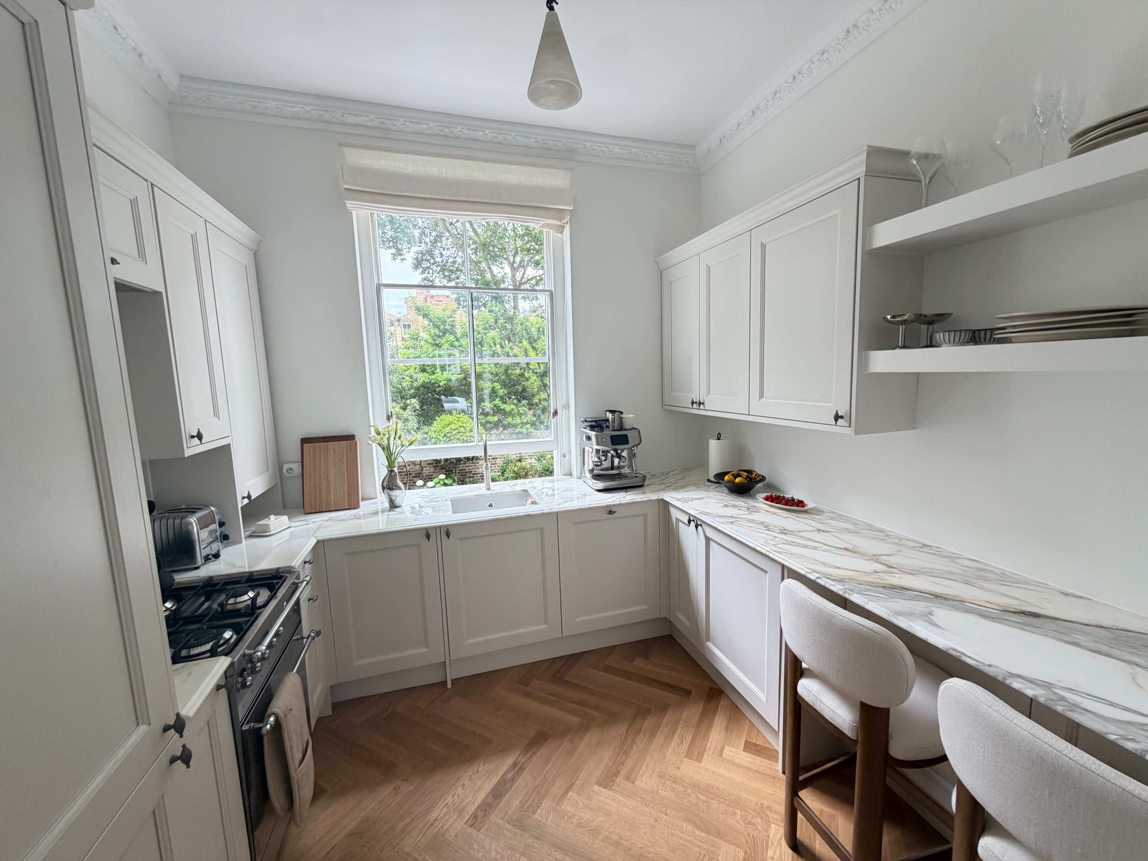 The image shows a modern kitchen with white cabinetry, a marble countertop, and a large window overlooking a garden.