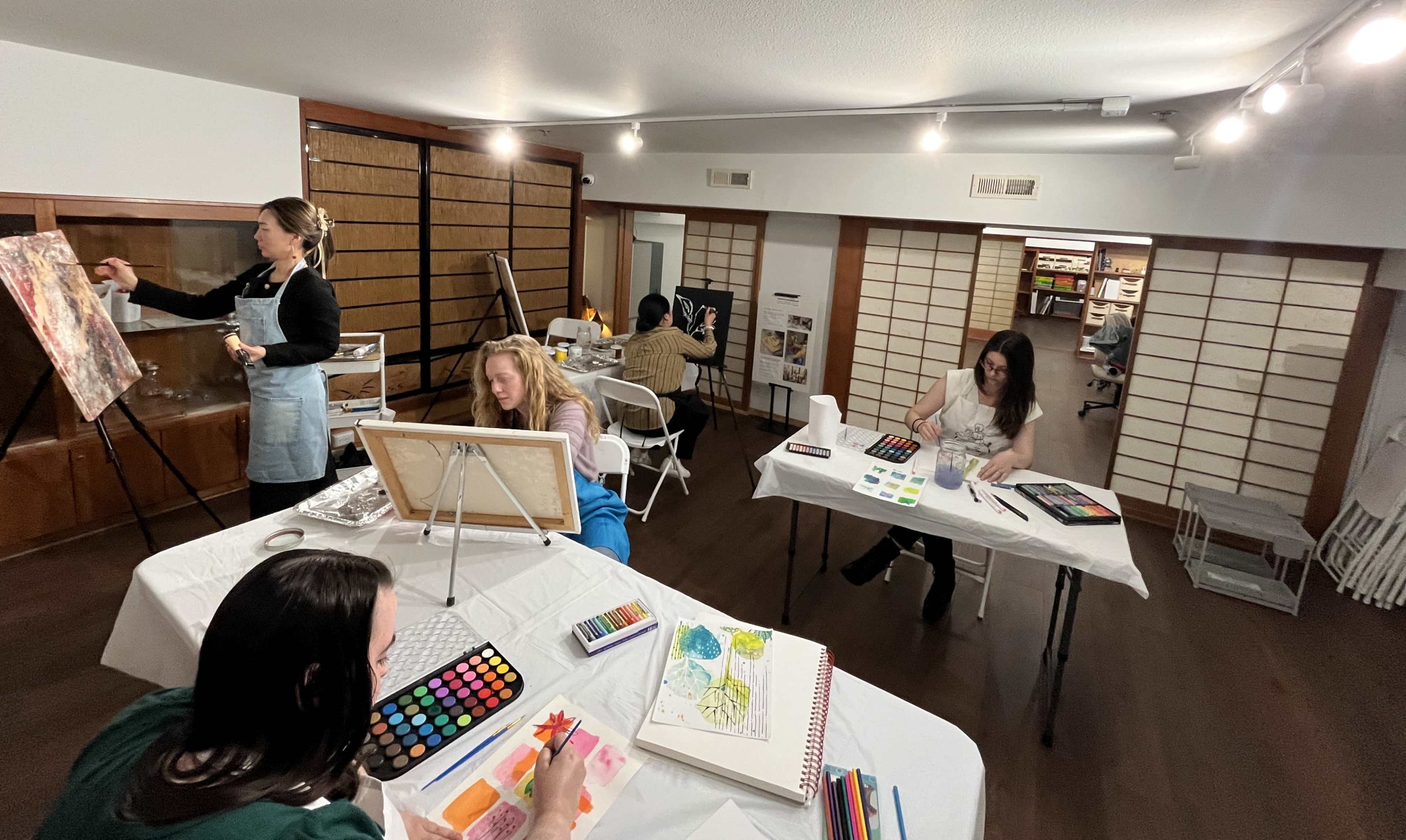 The image shows a spacious art studio with several people engaged in painting and drawing activities at tables, while one individual works on a canvas on an easel.