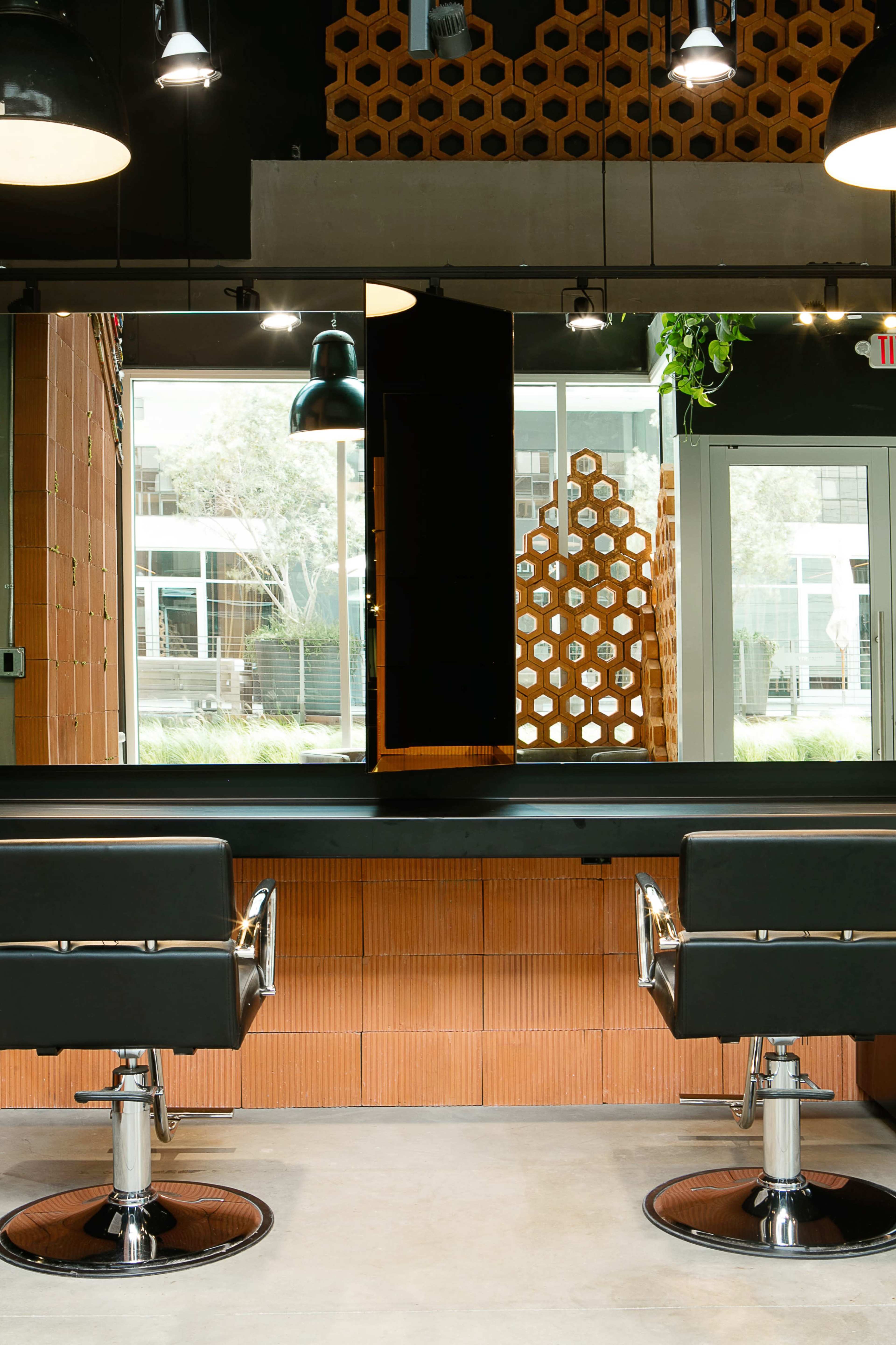 A modern hair salon features two black styling chairs facing a large mirror against a backdrop of geometric wooden panels and glass doors.