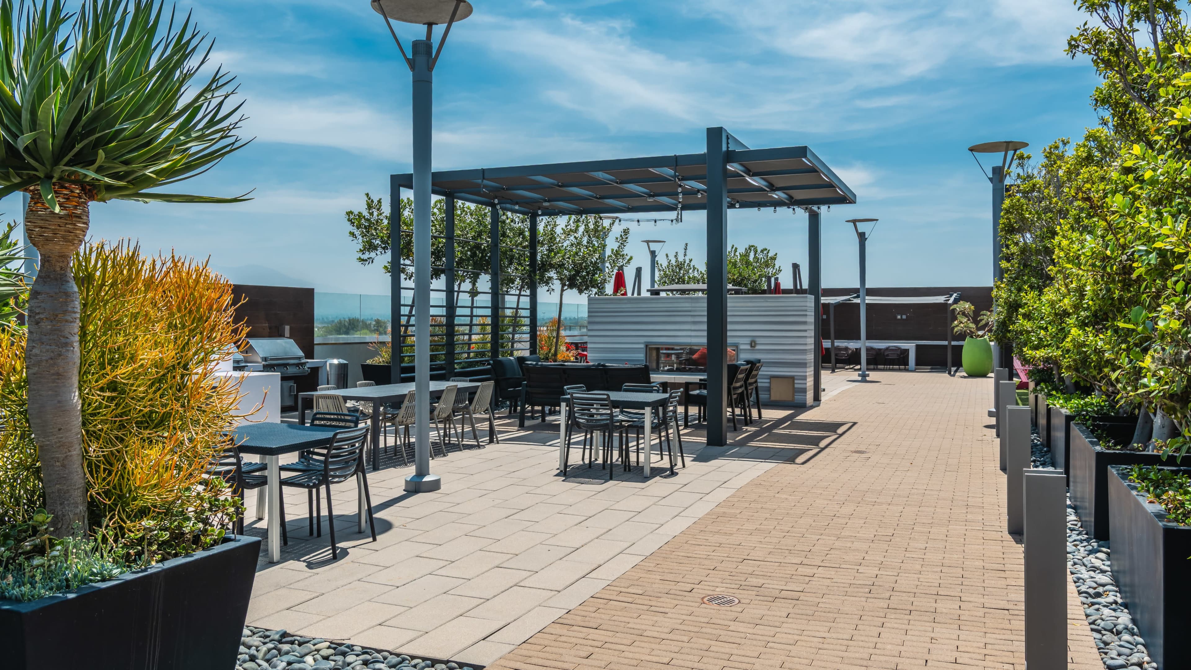 The image shows a rooftop terrace with metal seating, a grill area, and landscaped greenery under a clear blue sky.
