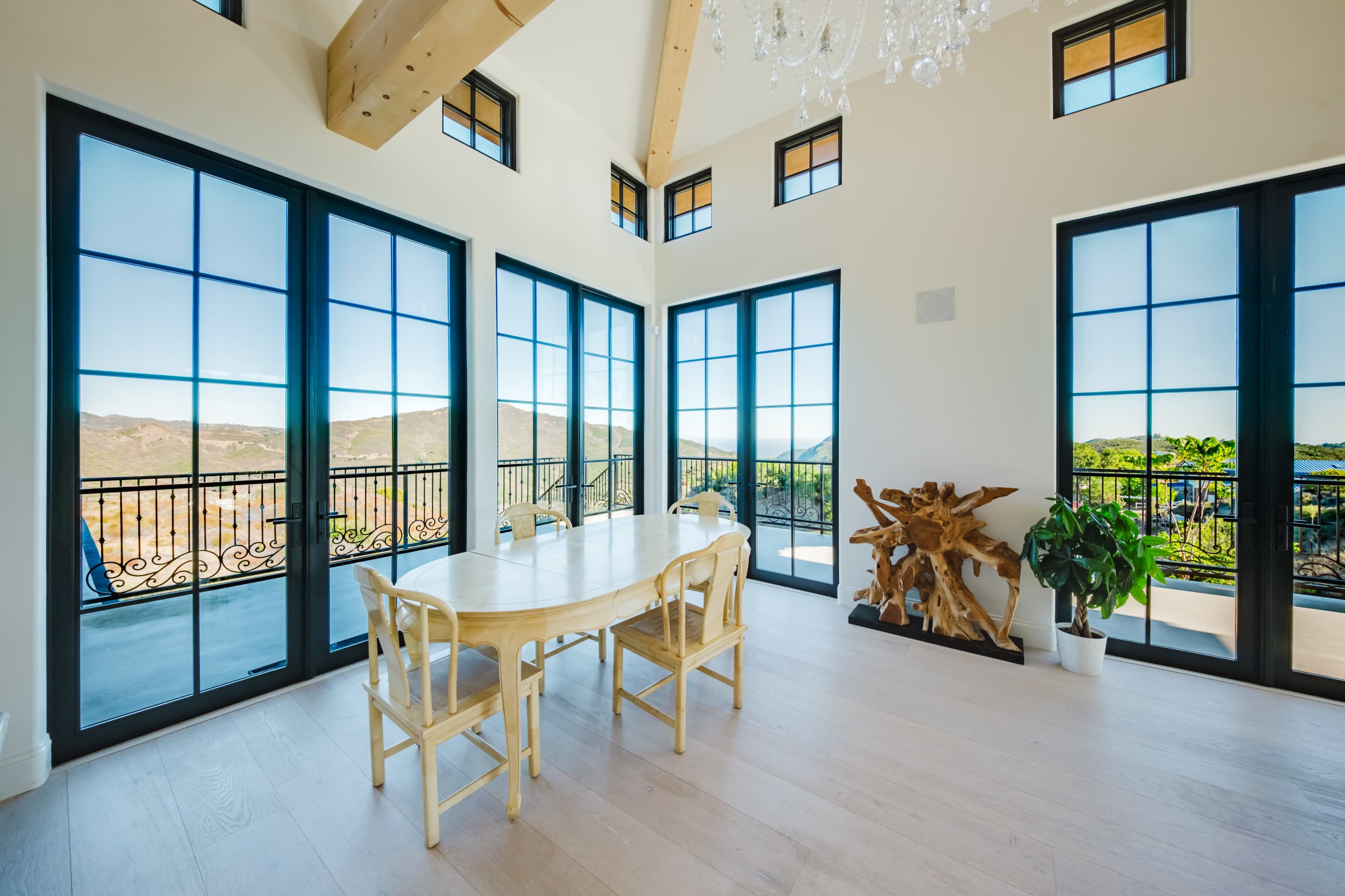 The image shows a bright dining area featuring a wooden table and chairs, surrounded by large windows with a view of a mountainous landscape.