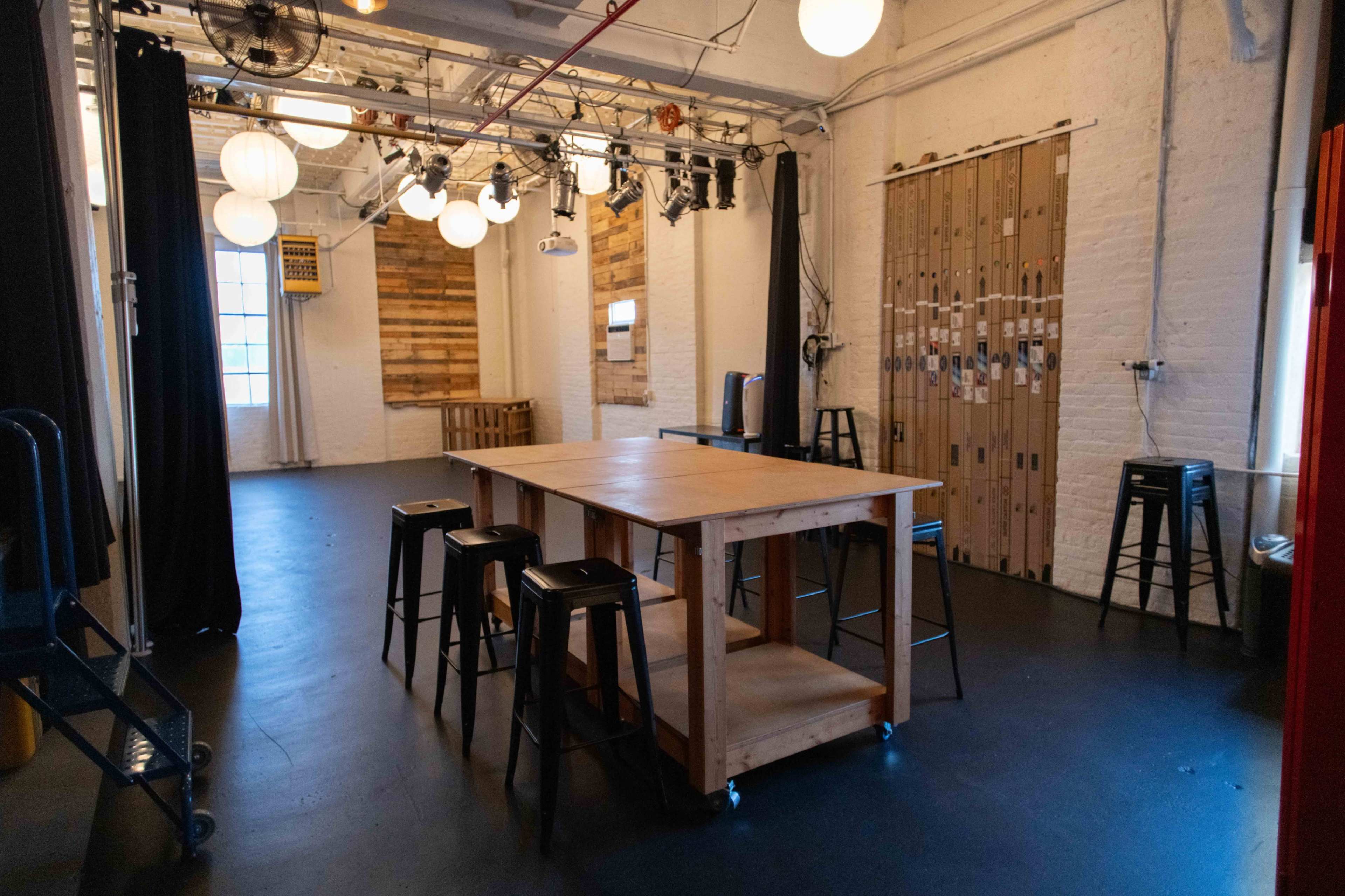 The image shows a spacious interior with a wooden table and several metal stools, illuminated by hanging light fixtures and featuring a backdrop of wooden panels.