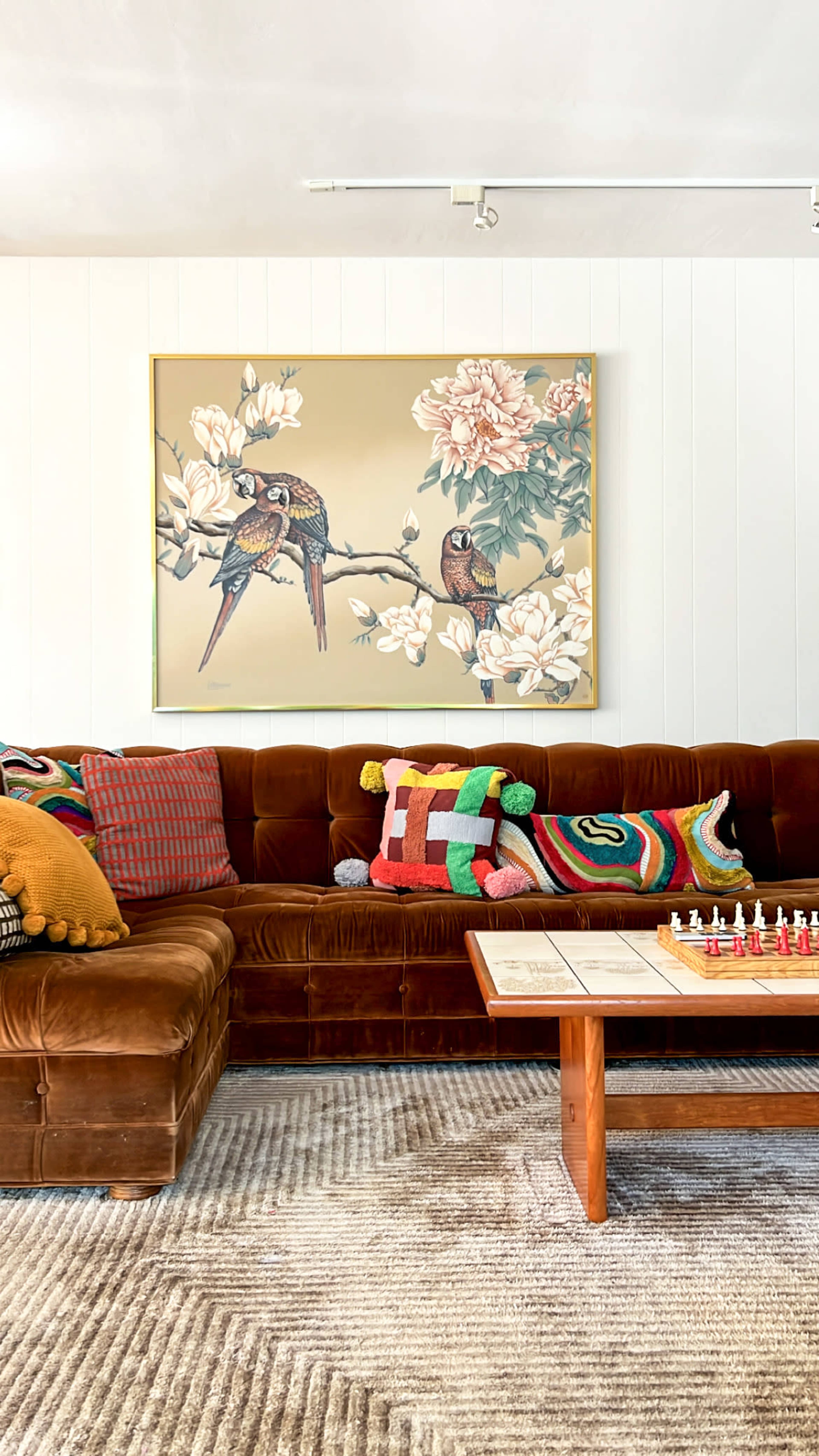 The image depicts a cozy living room with a brown velvet sectional sofa adorned with colorful pillows, a coffee table featuring a chessboard, and a large decorative painting of birds among flowers on the wall.