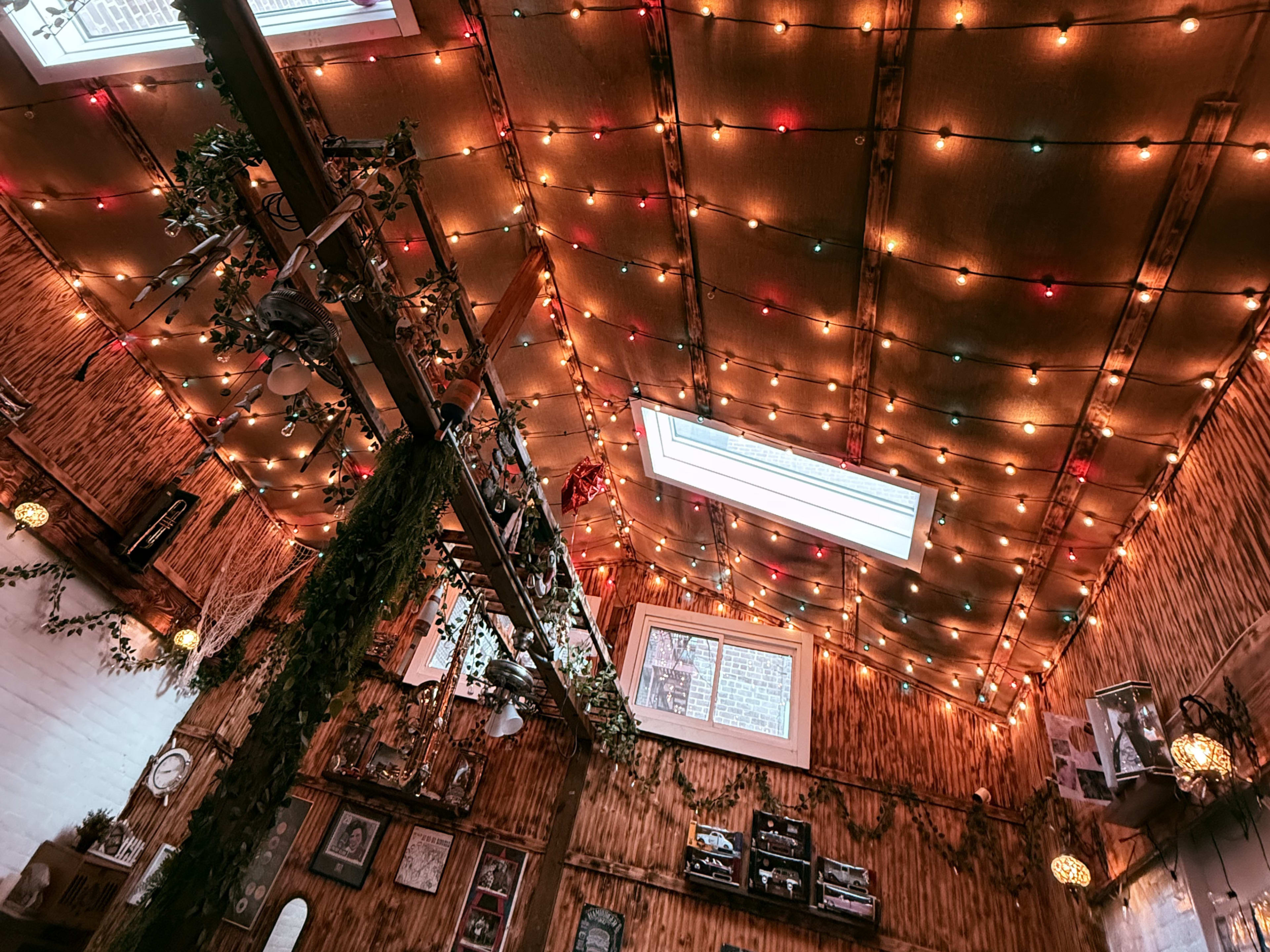 The image shows a cozy interior with a high ceiling adorned with colorful string lights and greenery, creating a warm ambiance.