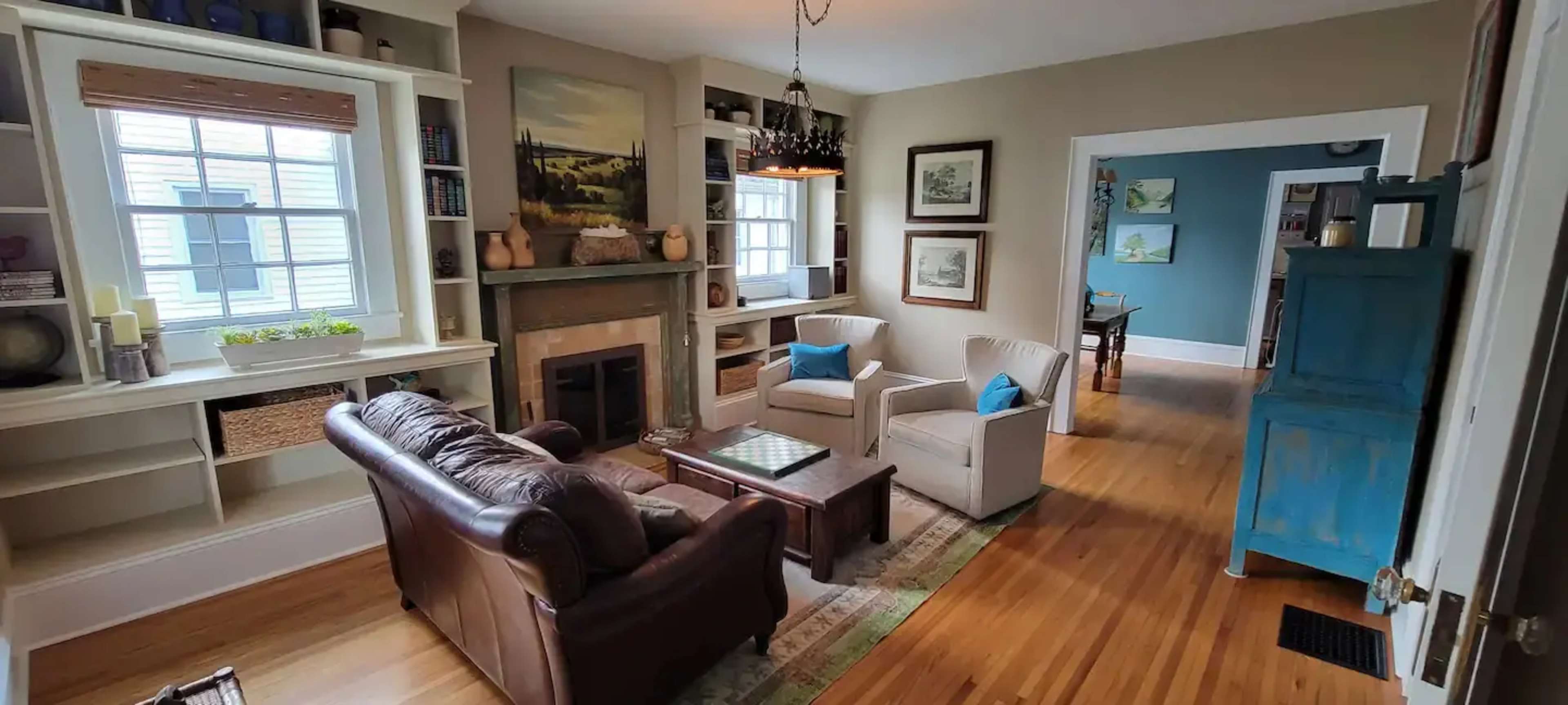 A comfortable living room features a leather sofa, two upholstered chairs, a coffee table, and a fireplace, with bookshelves and artwork on the walls.