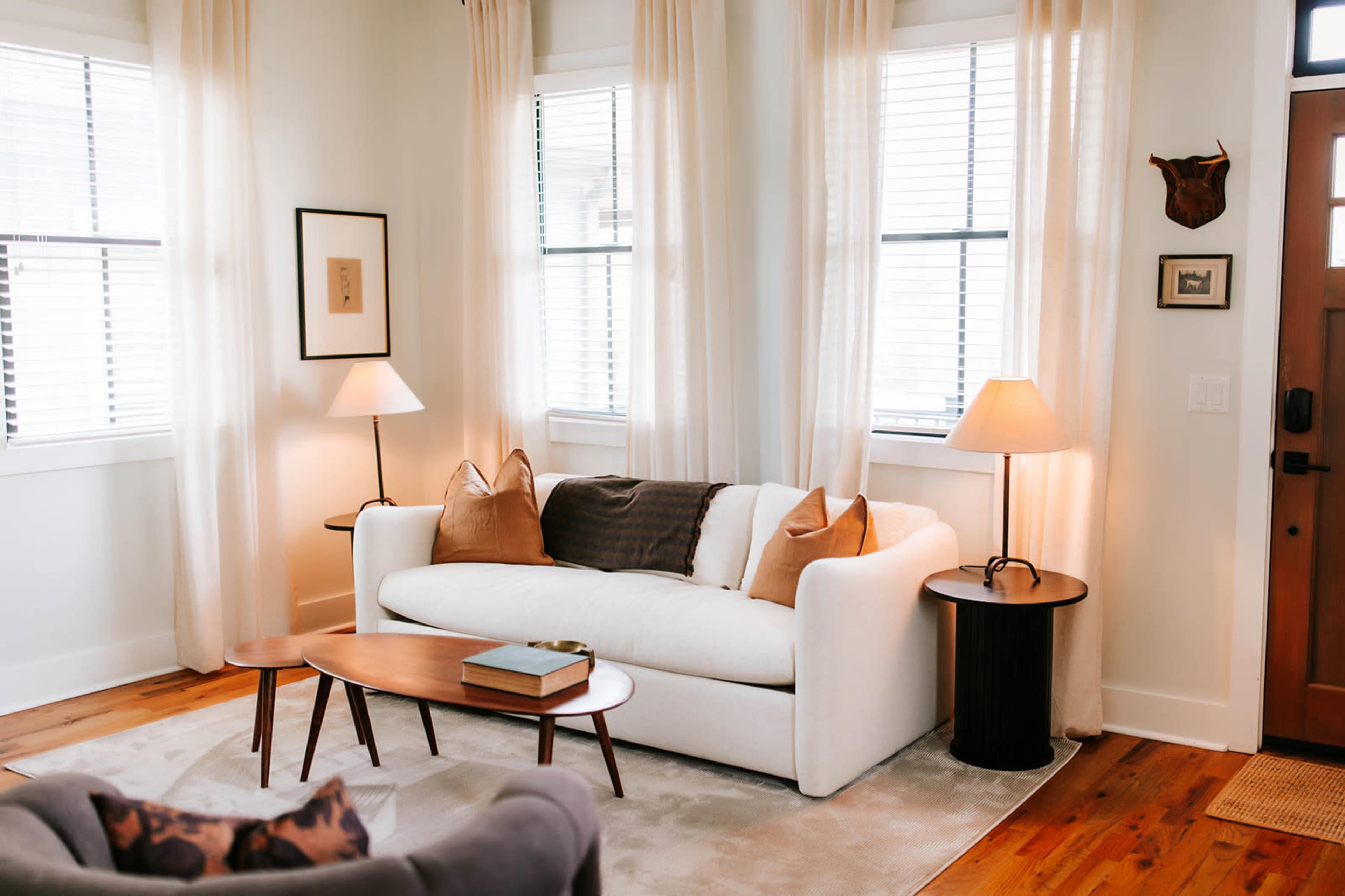The image depicts a cozy living room featuring a white sofa with decorative pillows, a wooden coffee table, and lamps illuminating the space, alongside large windows with sheer curtains.