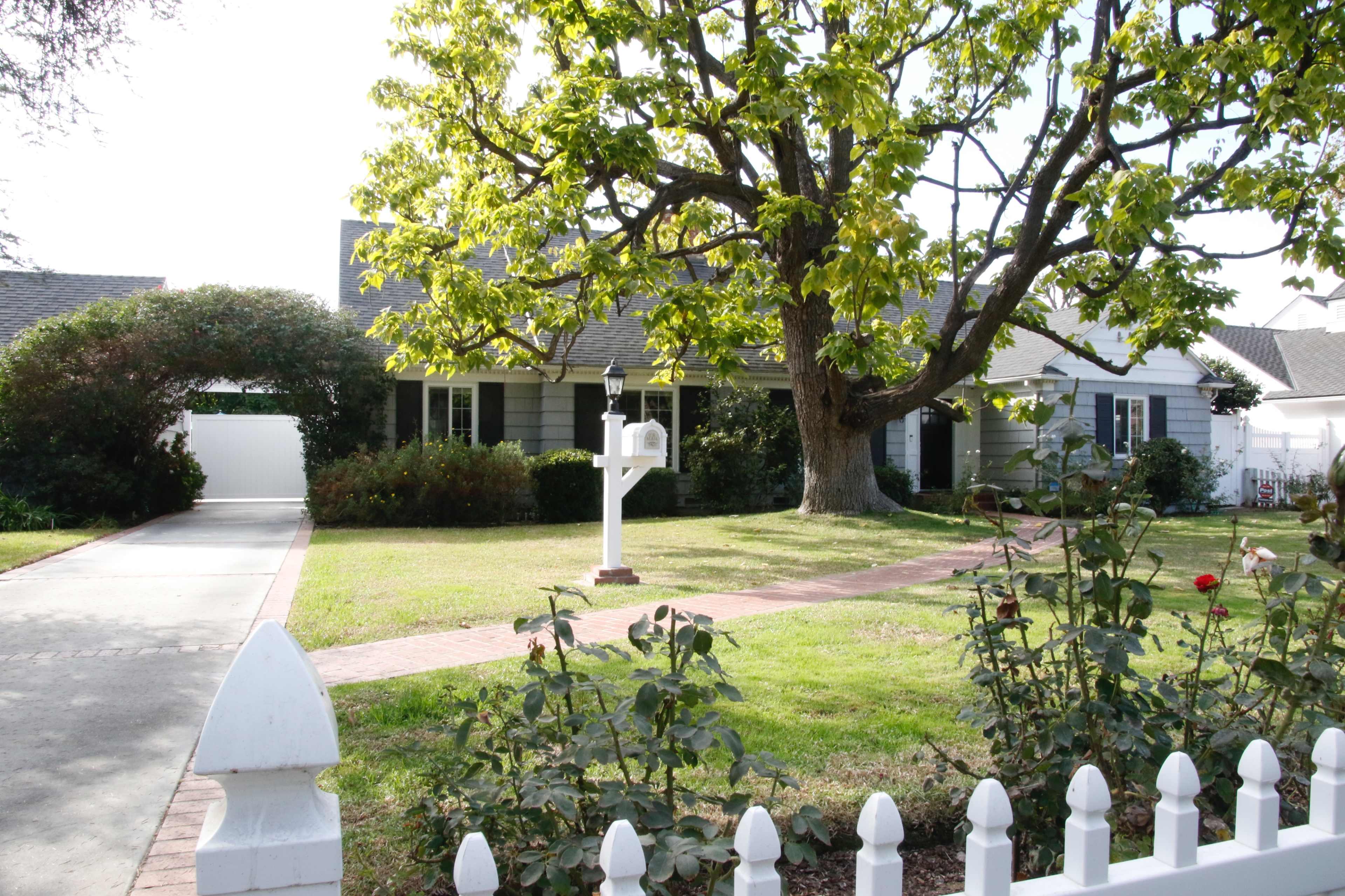 A single-story house with a porch is surrounded by a well-kept lawn, a white picket fence, and a large tree.
