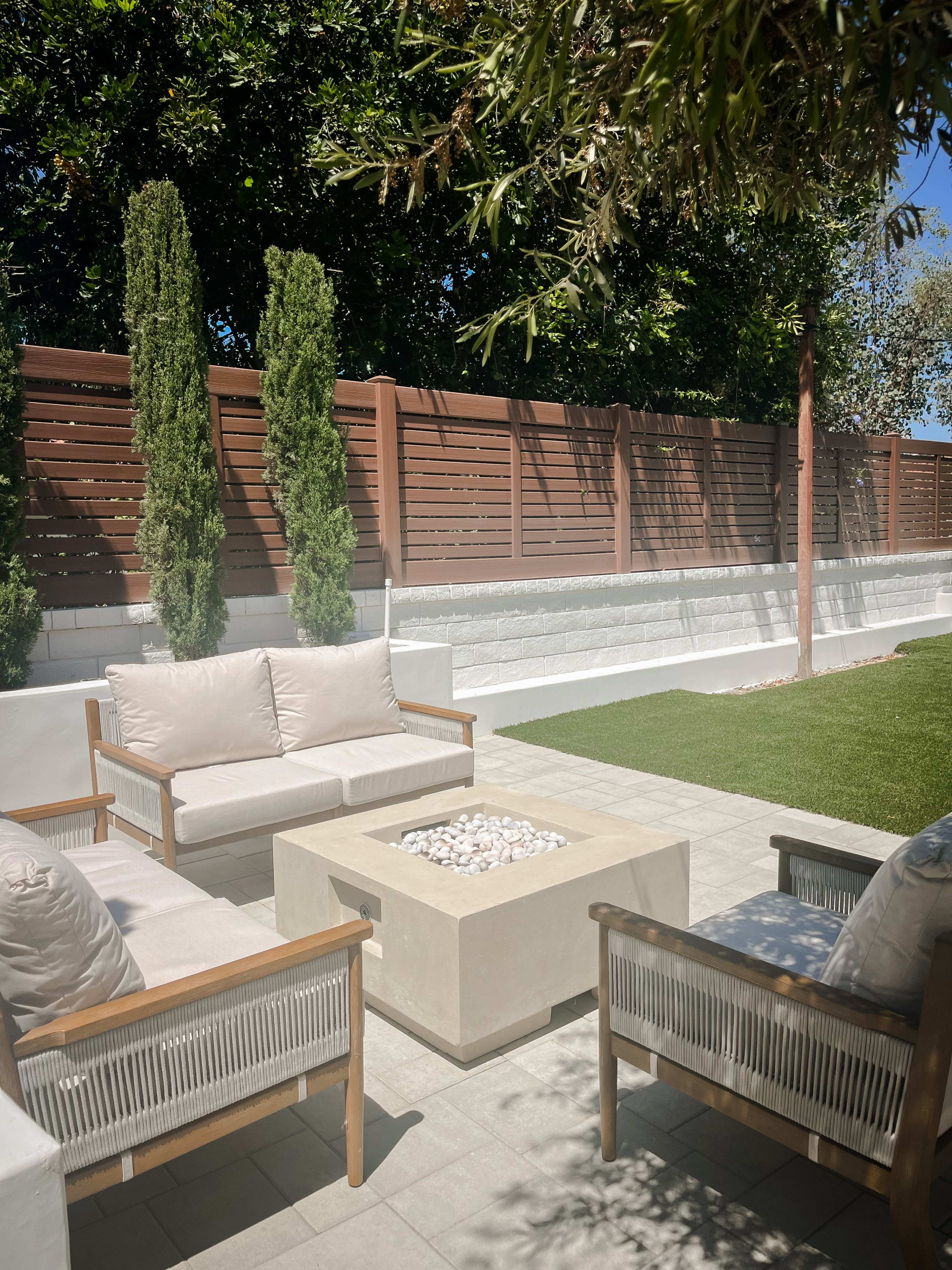 A patio area features two cushioned chairs flanking a square fire pit, with a fenced yard and greenery in the background.