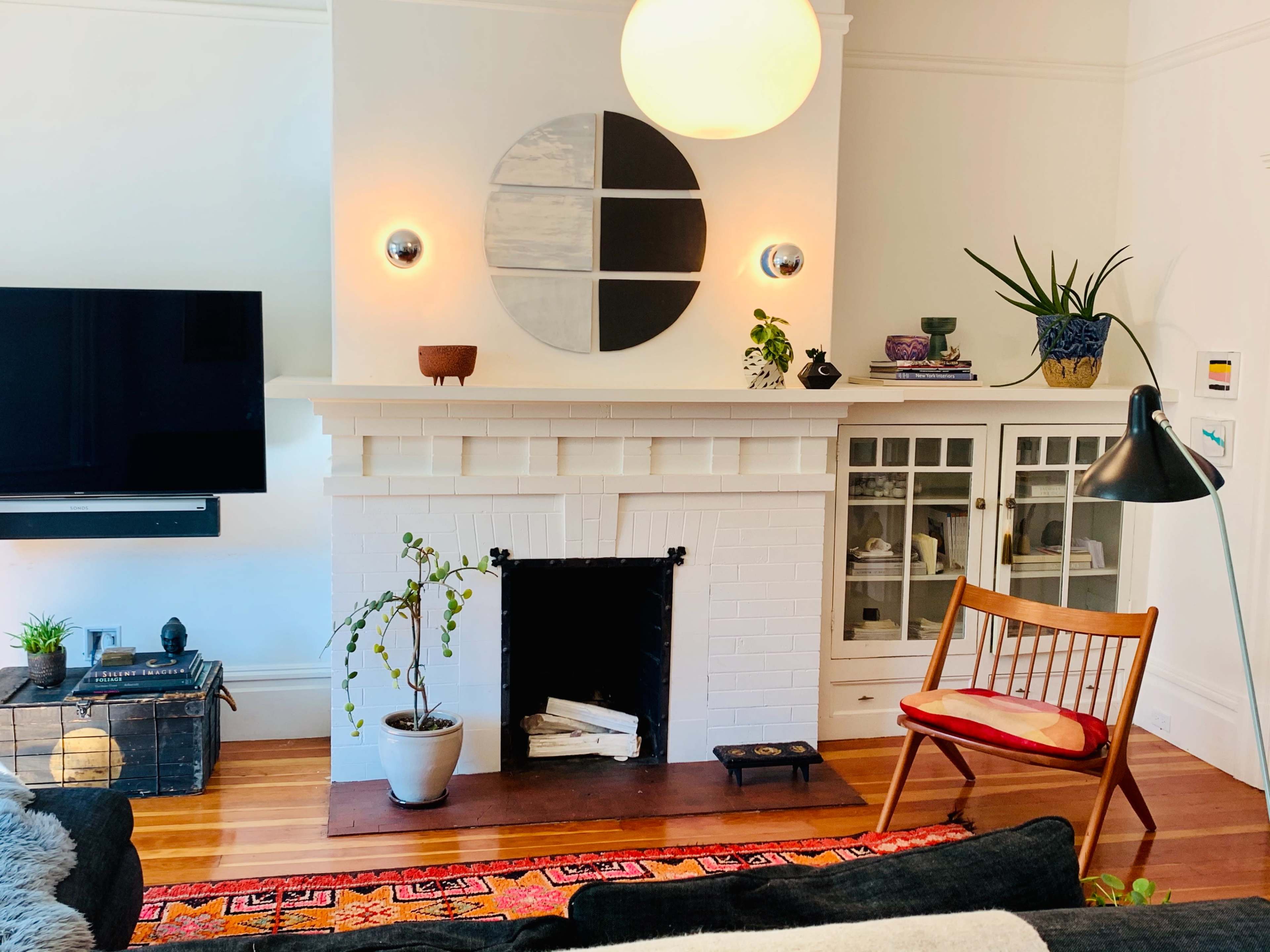 A living room with a white brick fireplace, a circular mirror above it, and a wooden chair beside a lamp, all complemented by a patterned rug.
