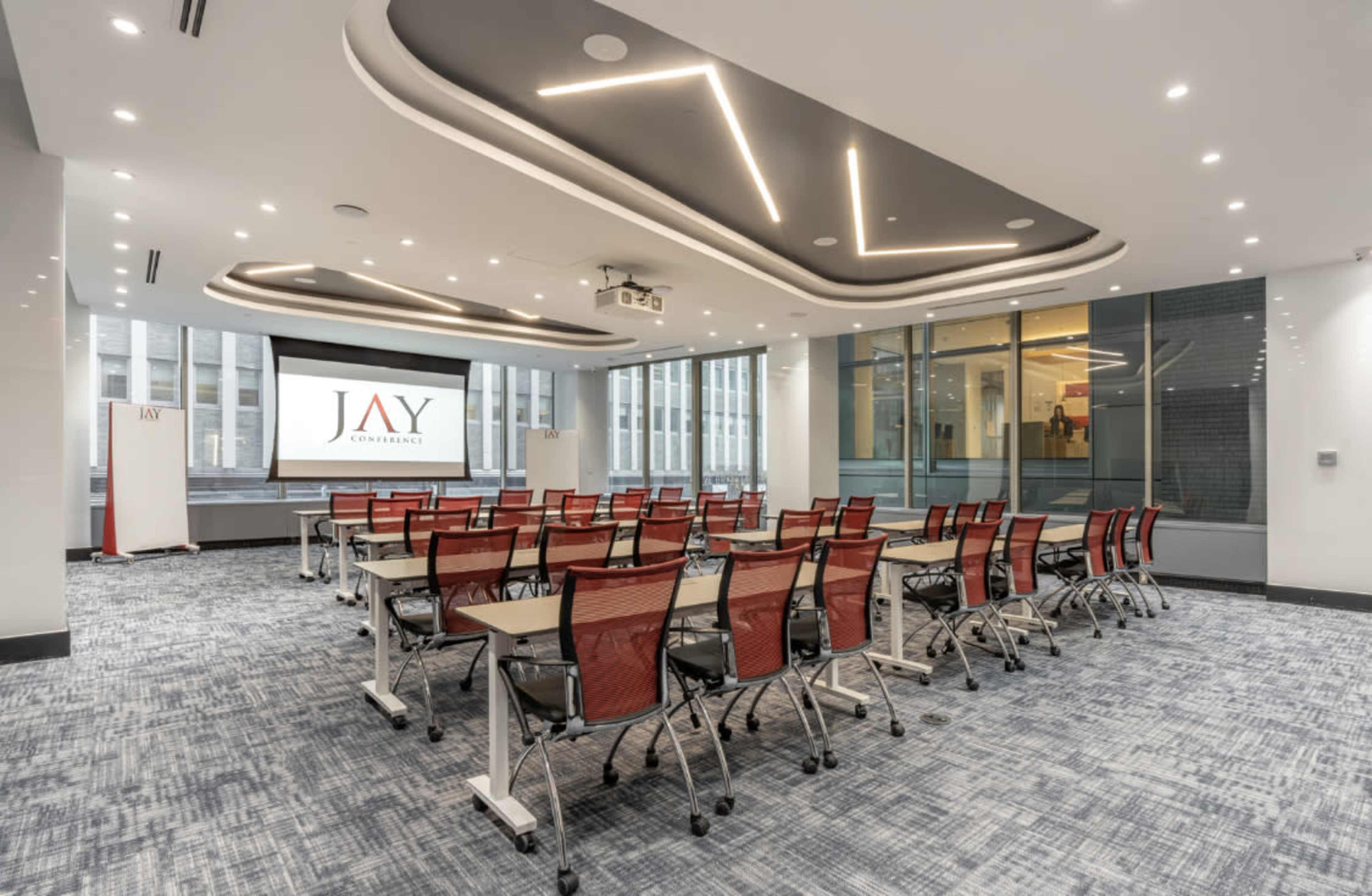 A modern conference room features rows of movable tables and red chairs, with a large screen displaying the logo "JAY" on the front wall.