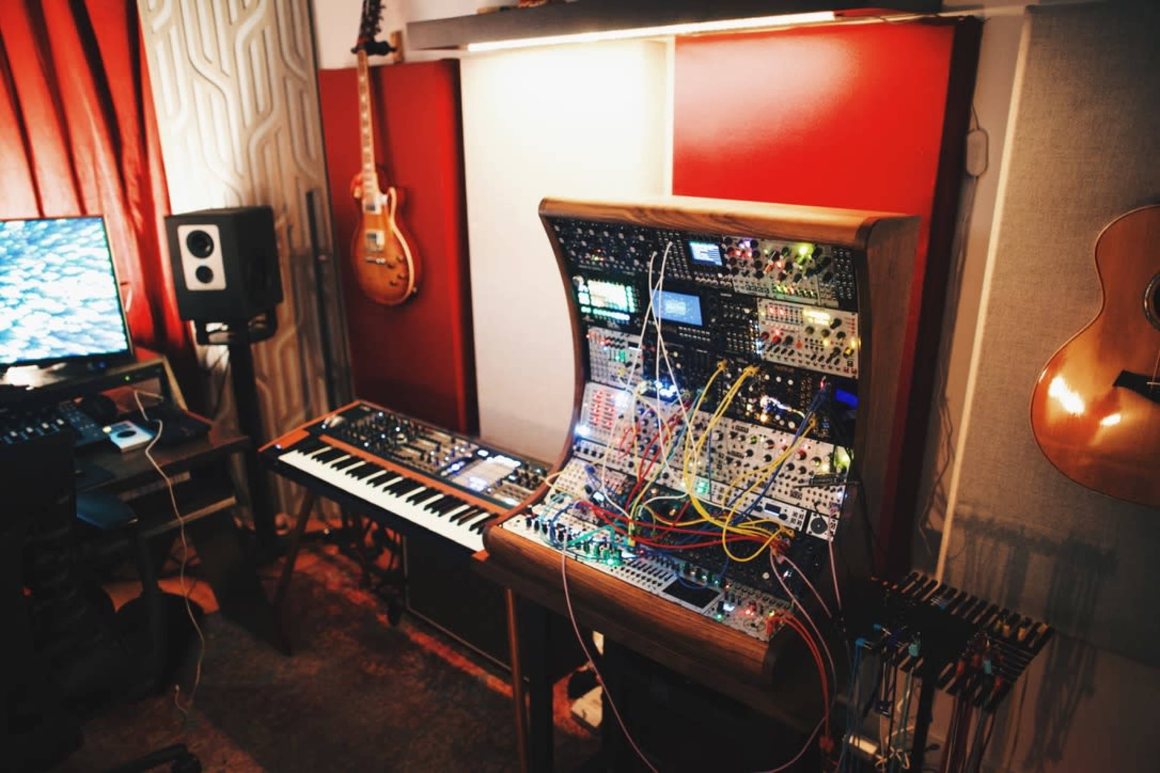 A music studio features a modular synthesizer setup with colorful wiring, a keyboard, and various audio equipment, surrounded by instruments on the walls.