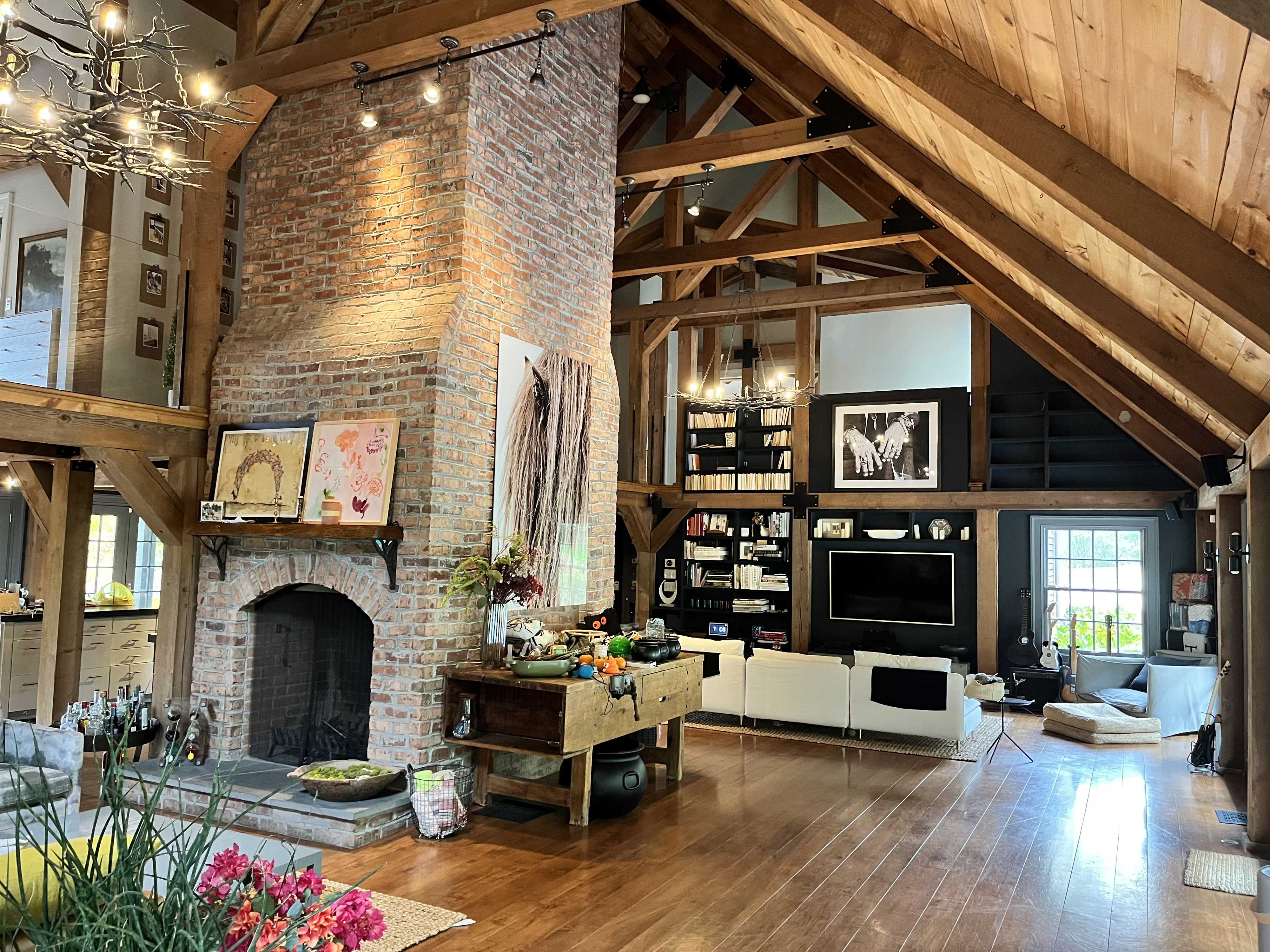 A spacious living room features a tall brick fireplace, wooden beams, and modern furnishings, illuminated by overhead lighting.