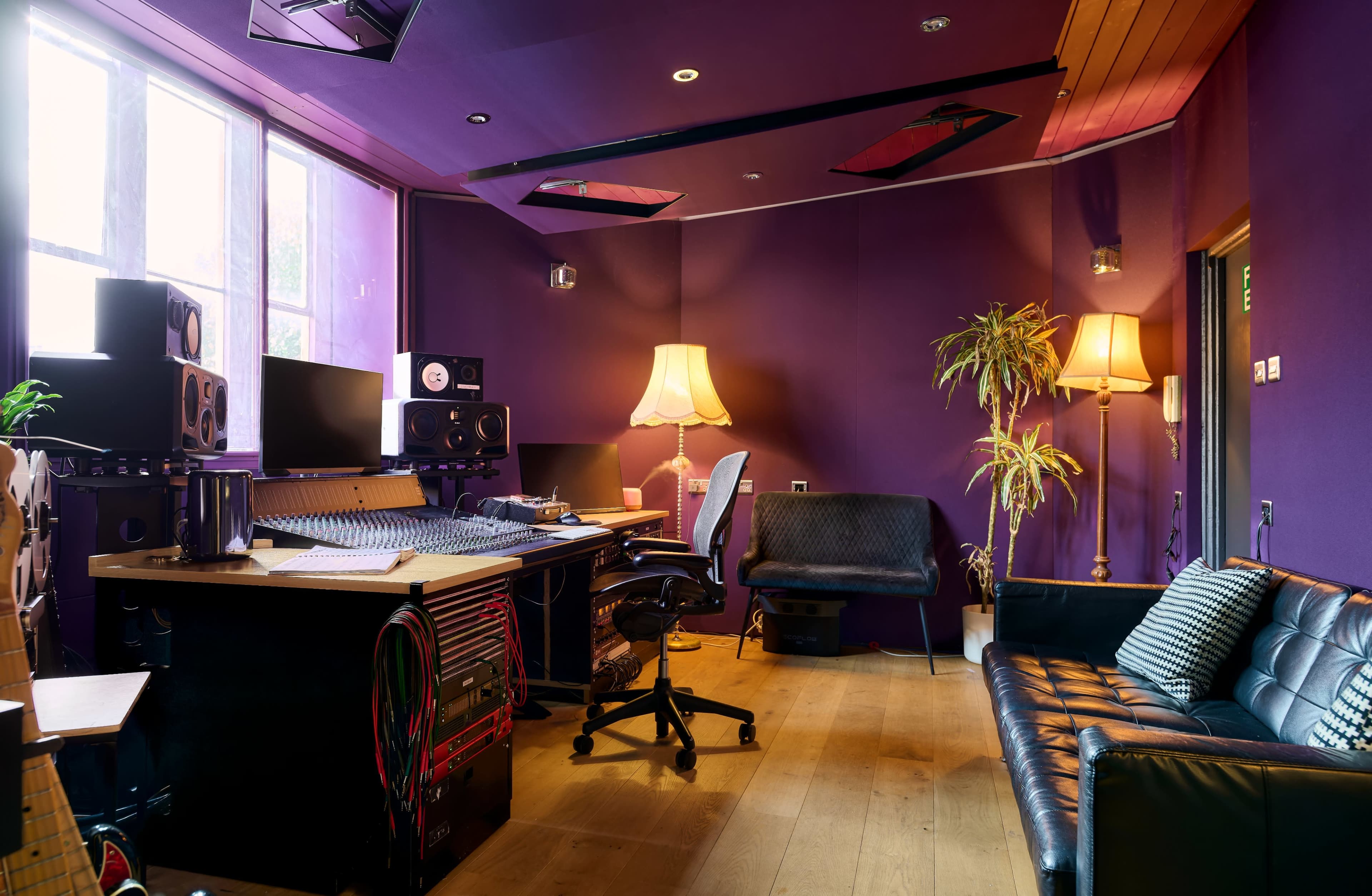 The image shows a recording studio with purple walls, various audio equipment, a desk with monitors, a sofa, and two lamps.