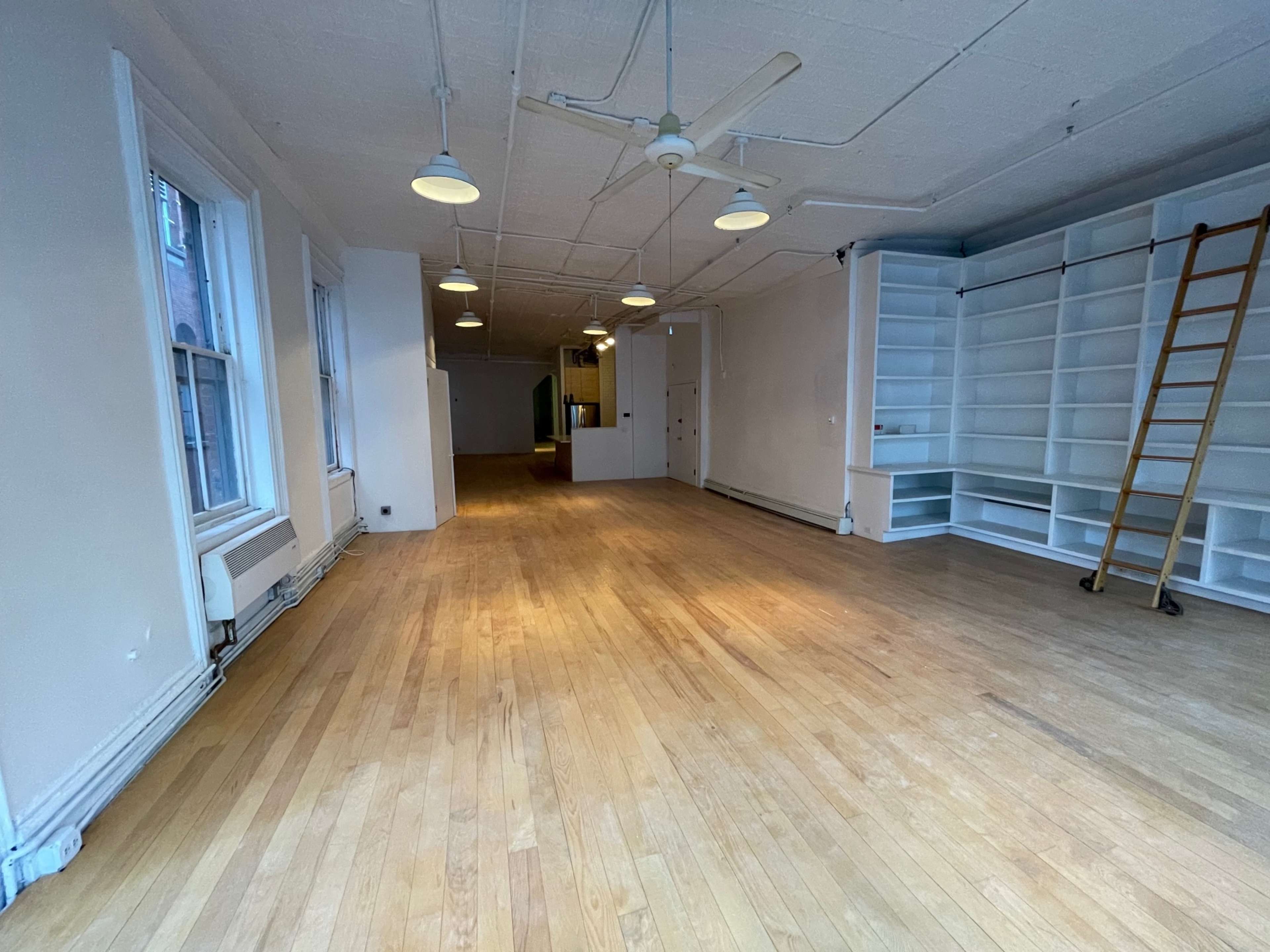 A spacious, empty room with wooden floors, white walls, and built-in shelves, illuminated by ceiling lights.