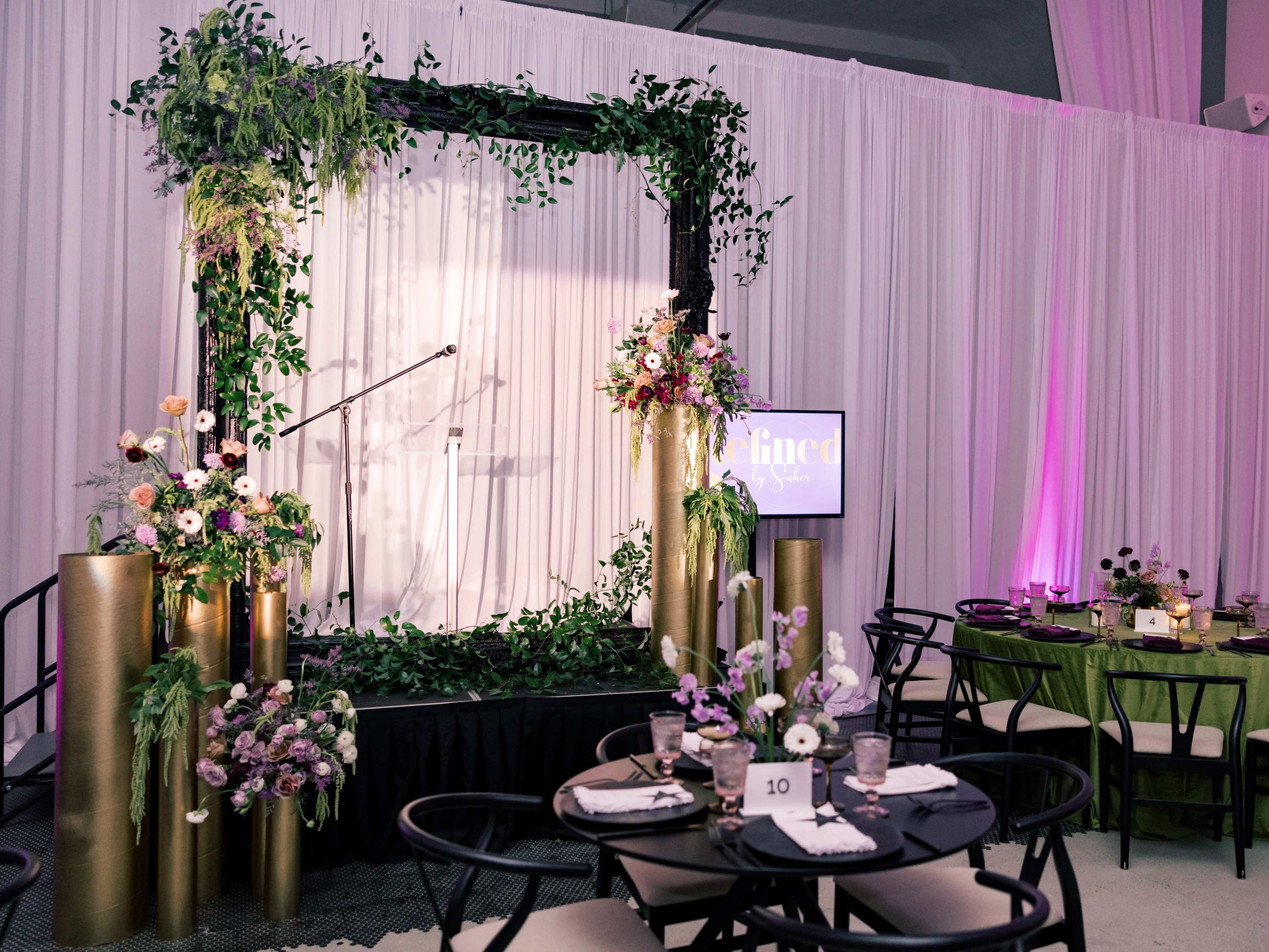 The image shows a decorated stage with a floral backdrop and a microphone, surrounded by tables set for an event with black chairs and floral centerpieces.