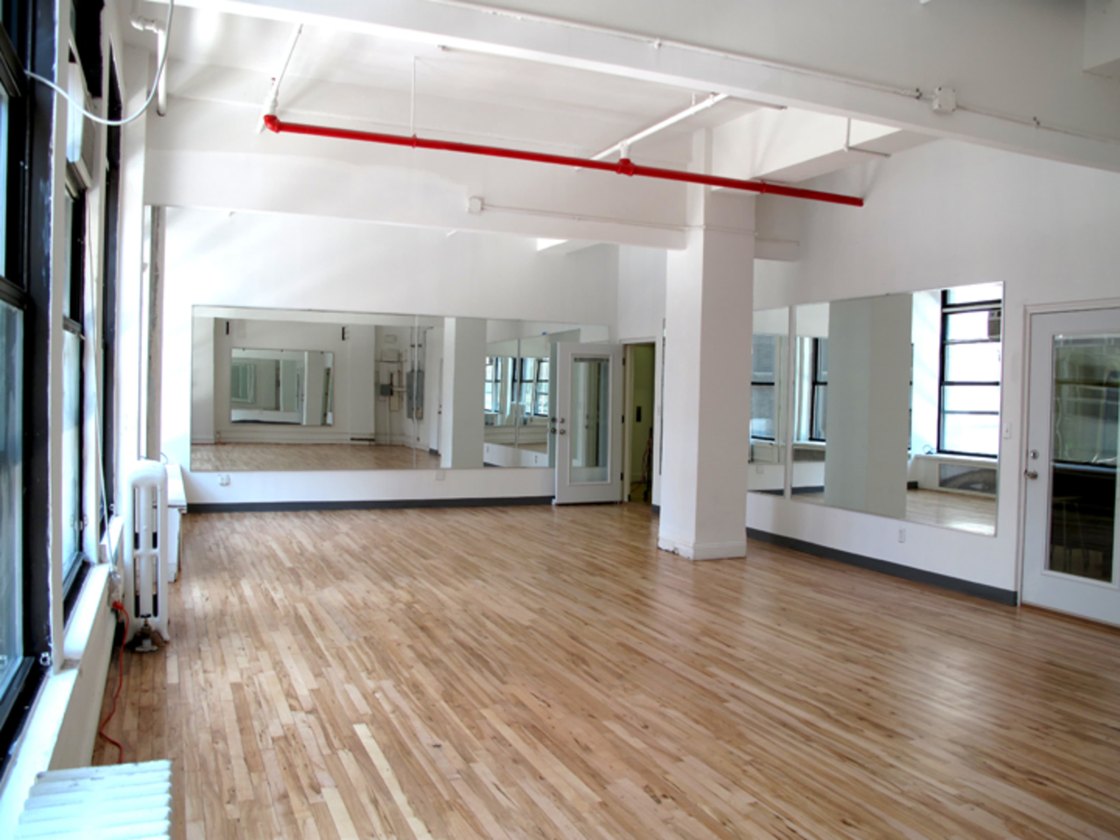 The image shows a spacious, empty room with wooden floors, large windows, and two mirrored walls.