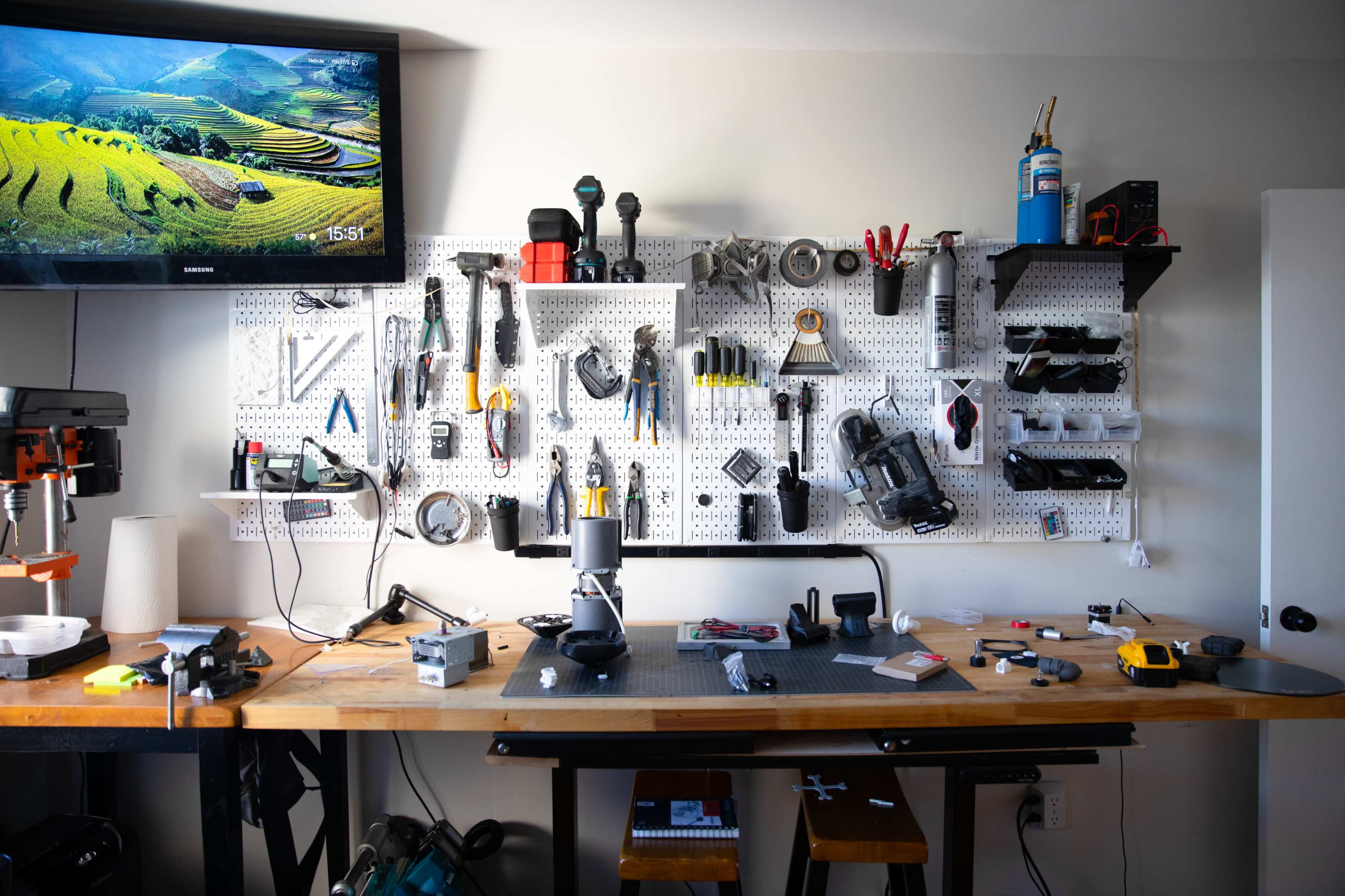 A workshop is shown with a wall-mounted pegboard displaying various tools and equipment, alongside a workspace featuring an array of instruments and a TV mounted above.