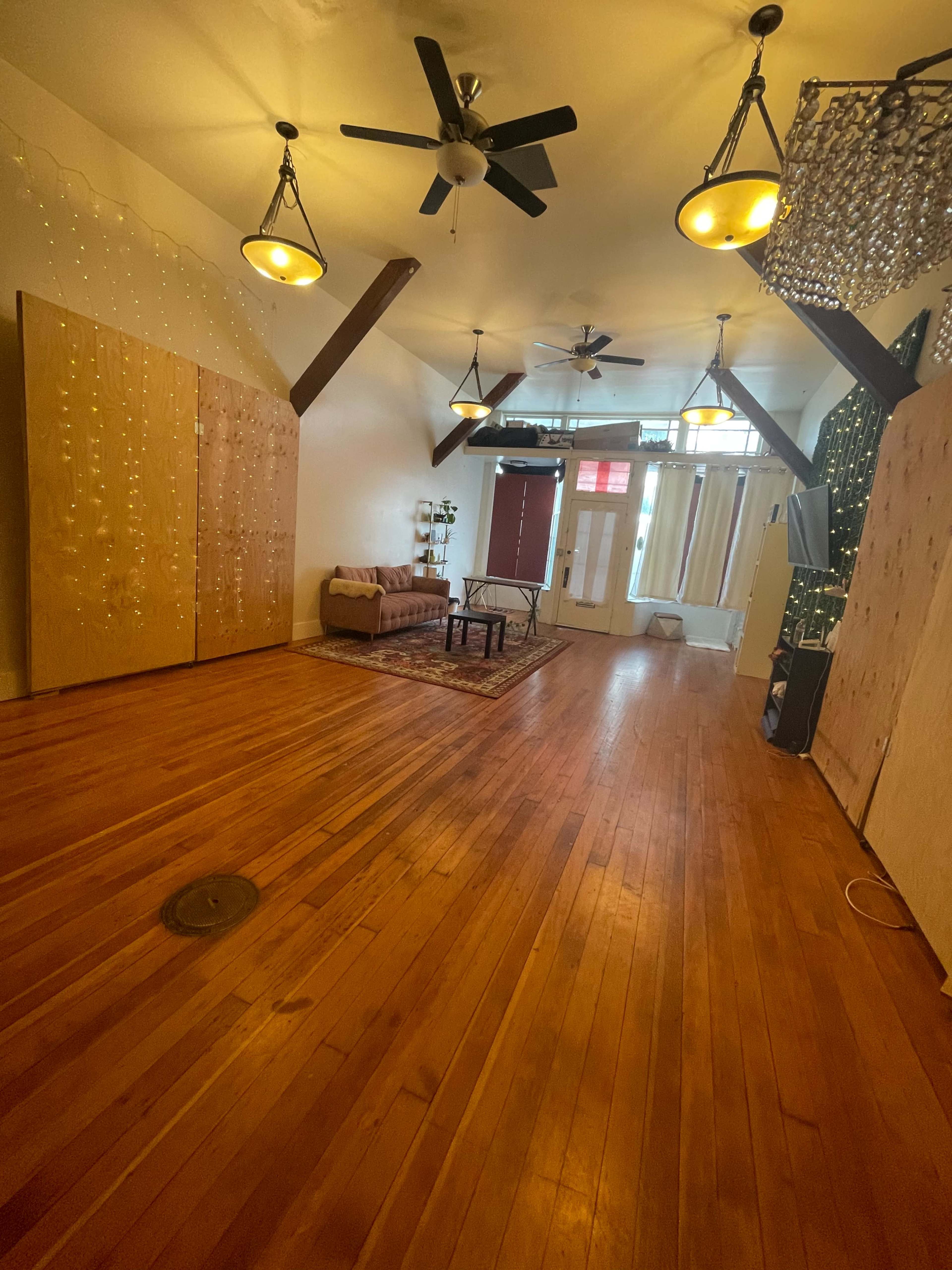 A spacious room with hardwood floors, large windows, and various lighting fixtures, featuring a couch and decorative elements along the walls.