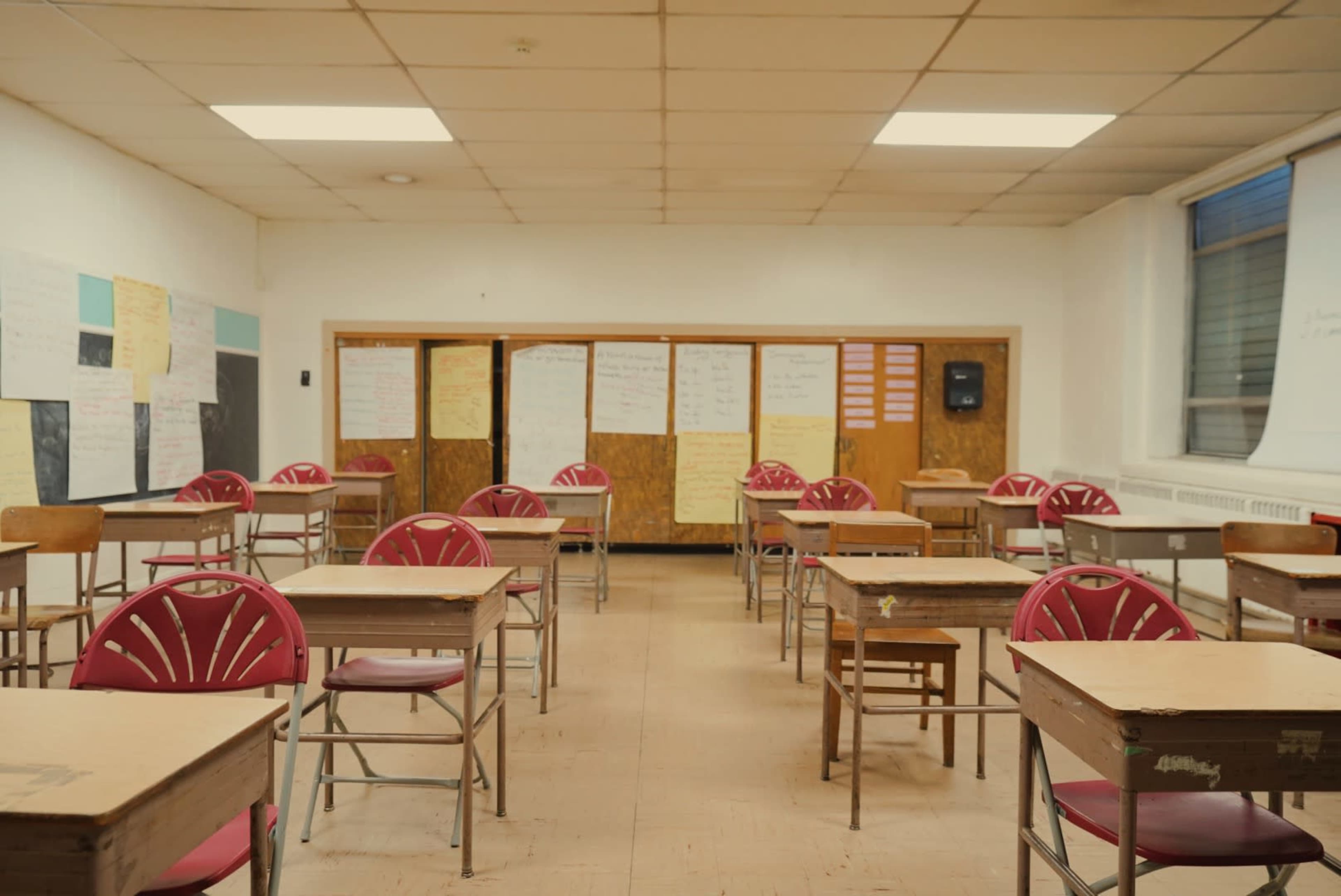 An empty classroom features wooden desks and chairs arranged in rows with paper-covered walls.