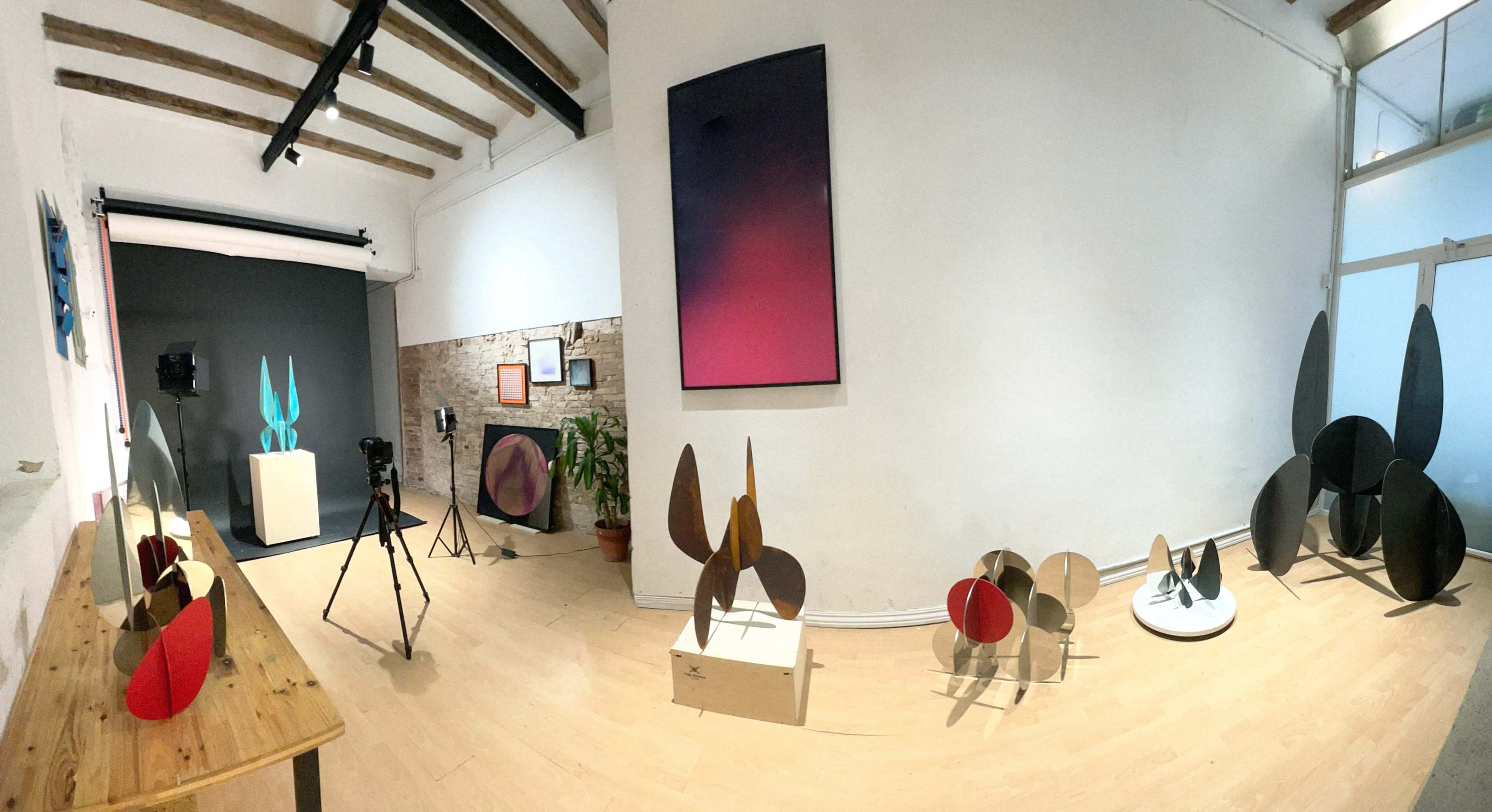 The image depicts an art gallery featuring various abstract sculptures arranged on a wooden table and the floor, with artwork displayed on the walls.