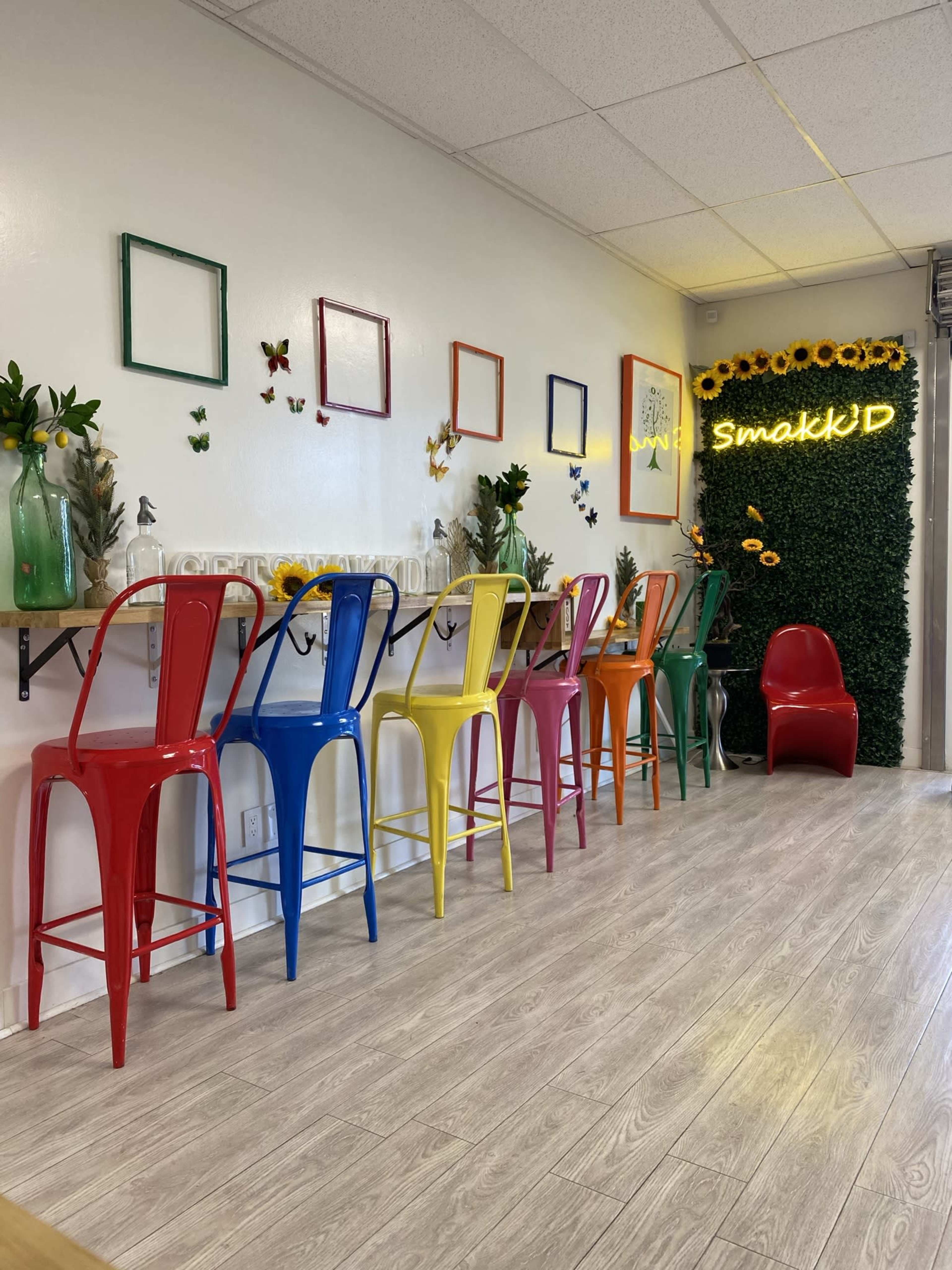 A brightly colored dining area with metal chairs in red, blue, yellow, and green arranged around a long wooden table, decorated with plants and wall art.