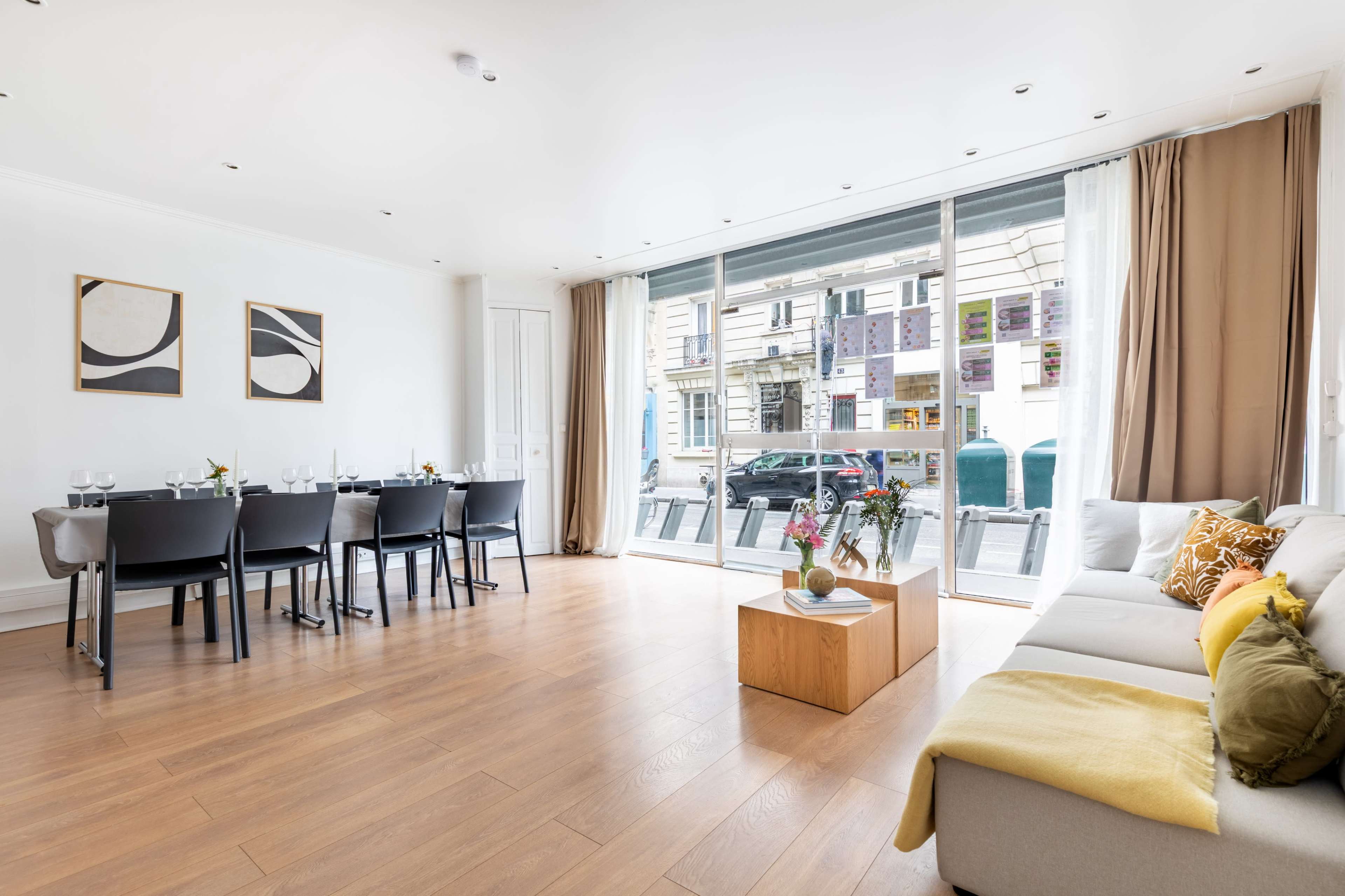 The image shows a modern, spacious living area with a dining table, a sofa, and large windows facing an outdoor street view.