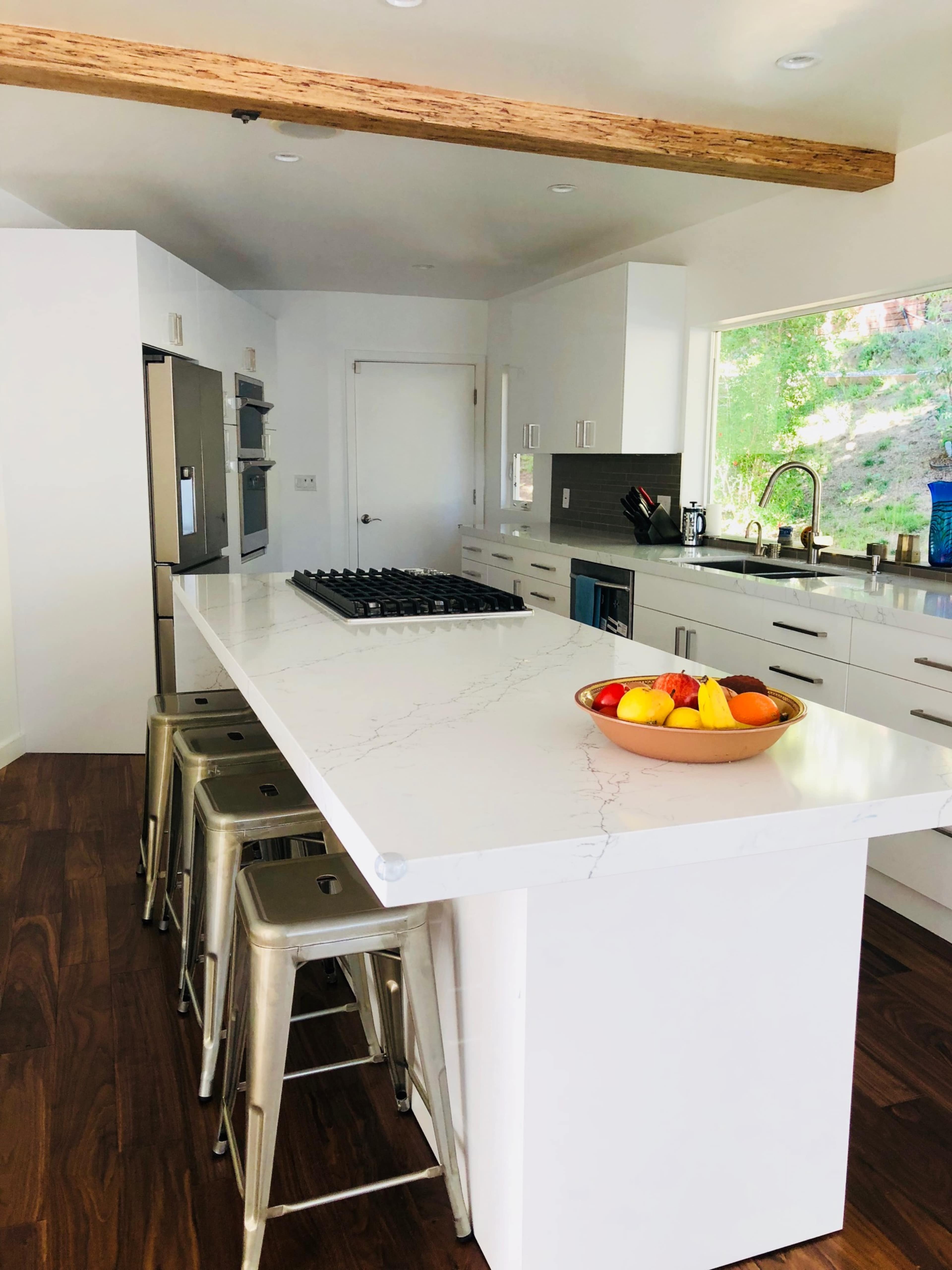 A modern kitchen features a large white island with a marble top and metal bar stools, surrounded by sleek cabinetry and a view of greenery outside.