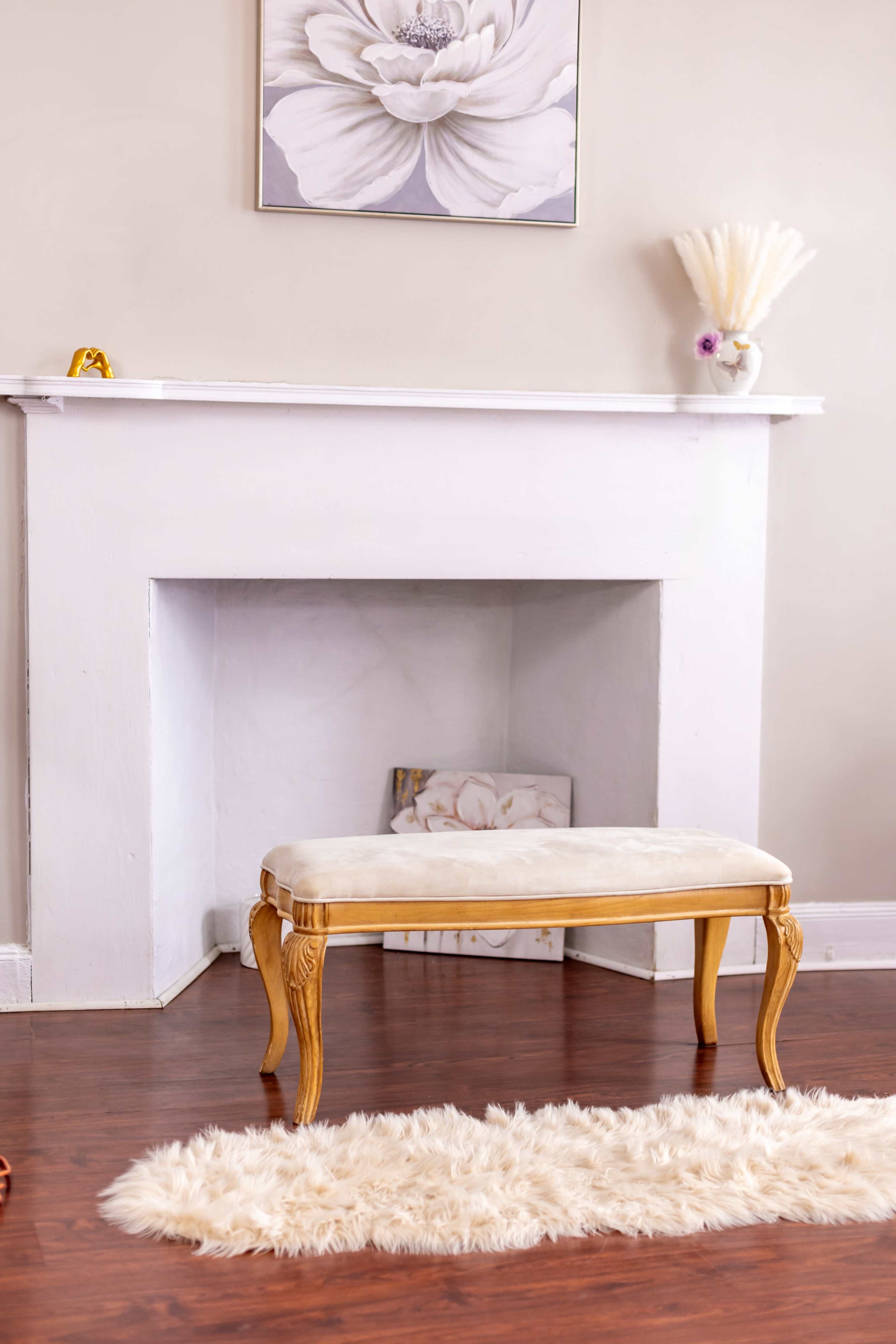 A wooden fireplace with a white mantel is positioned against a wall, featuring a decorative flower print, and a small, upholstered bench sits in front of it on a wooden floor covered by a plush rug.
