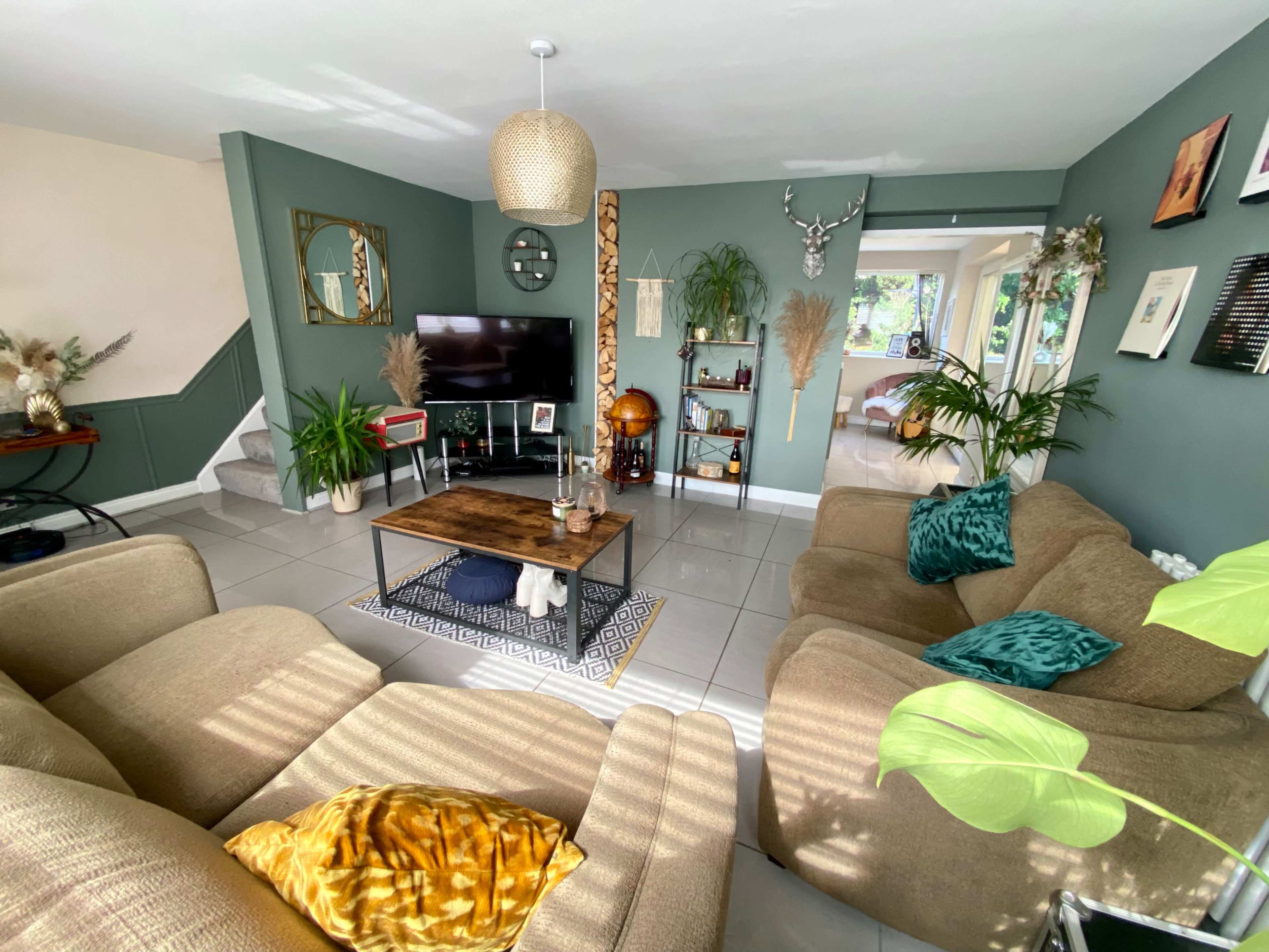 A cozy living room with beige sofas, a wooden coffee table, and various decorative plants, featuring a television on a wall surrounded by artwork.