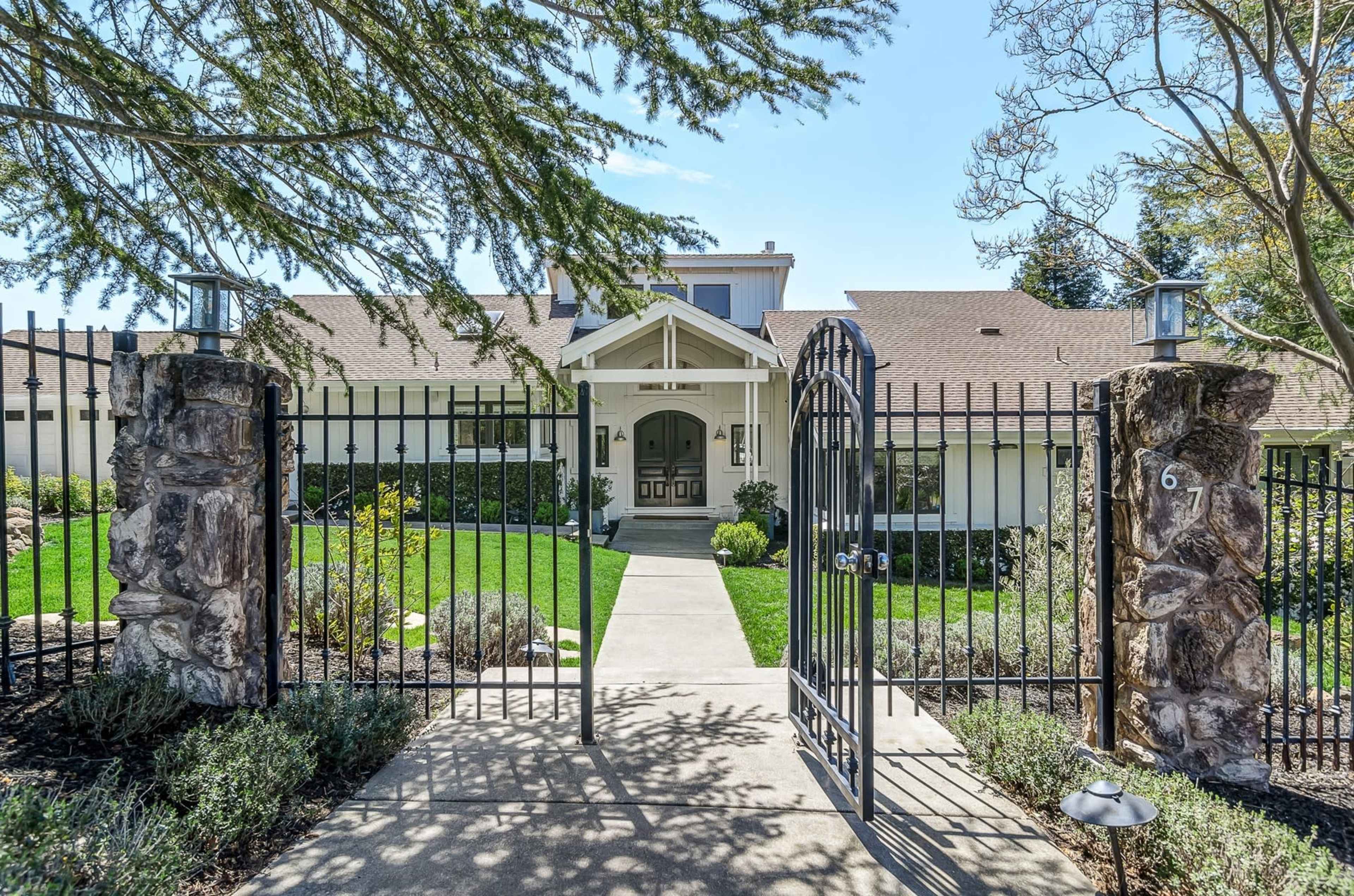 A stone gate opens to a well-maintained pathway leading to a modern house with a front porch and manicured landscaping.