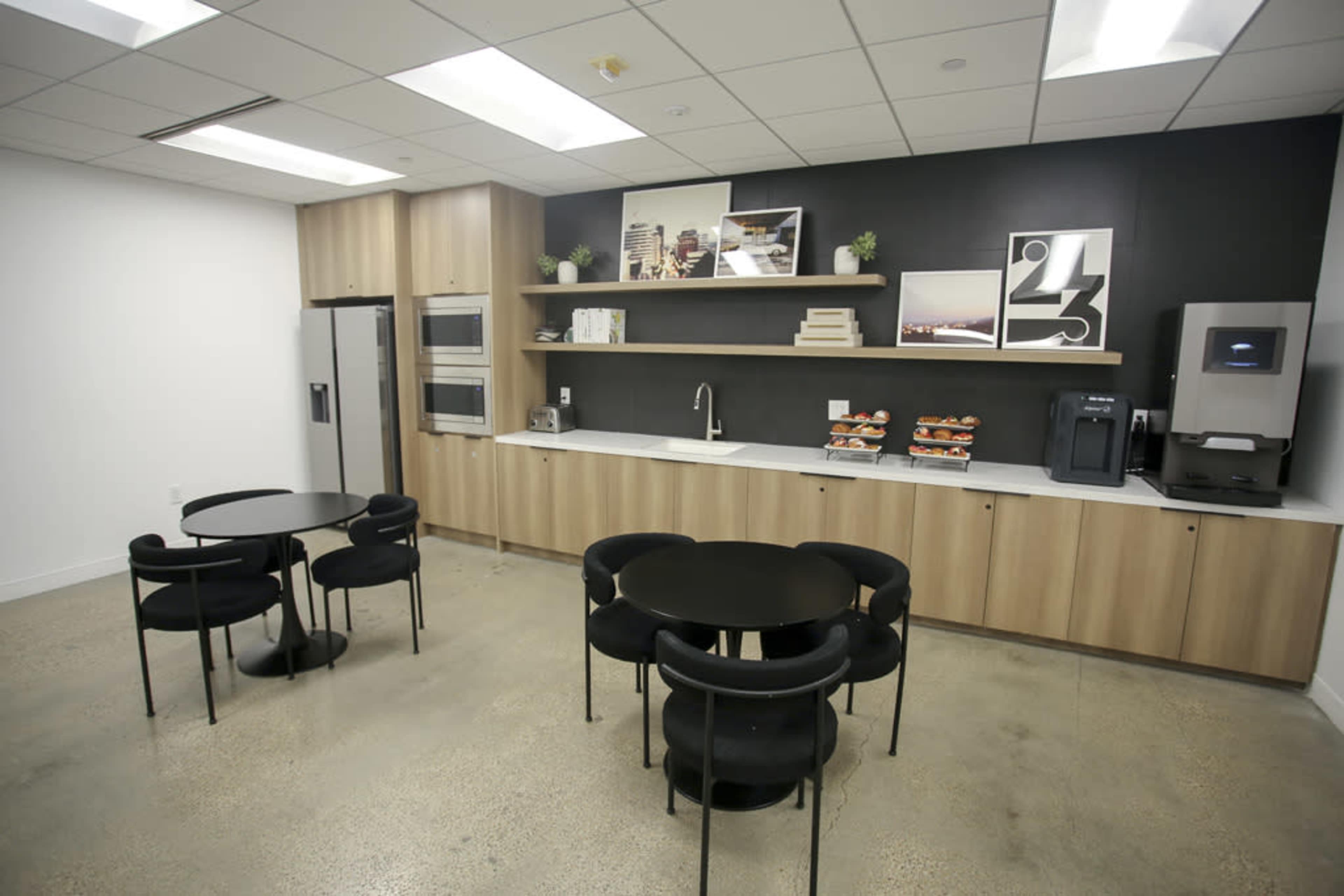 A modern break room with two circular black tables, a countertop with kitchen appliances, and a wall featuring decorative art pieces.