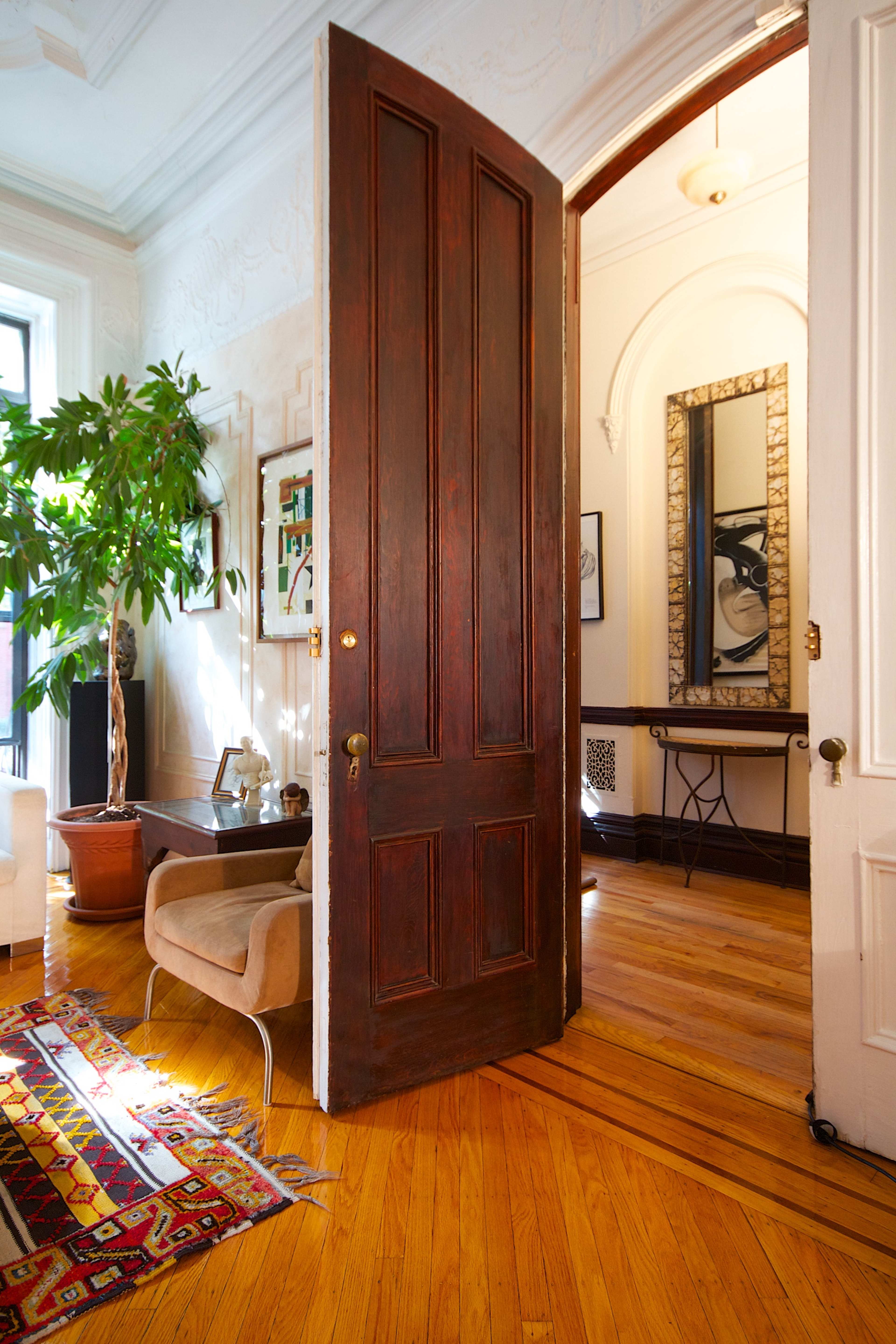 A partially open wooden door reveals a bright interior with a rug, a plant, and a large mirror on the opposite wall.
