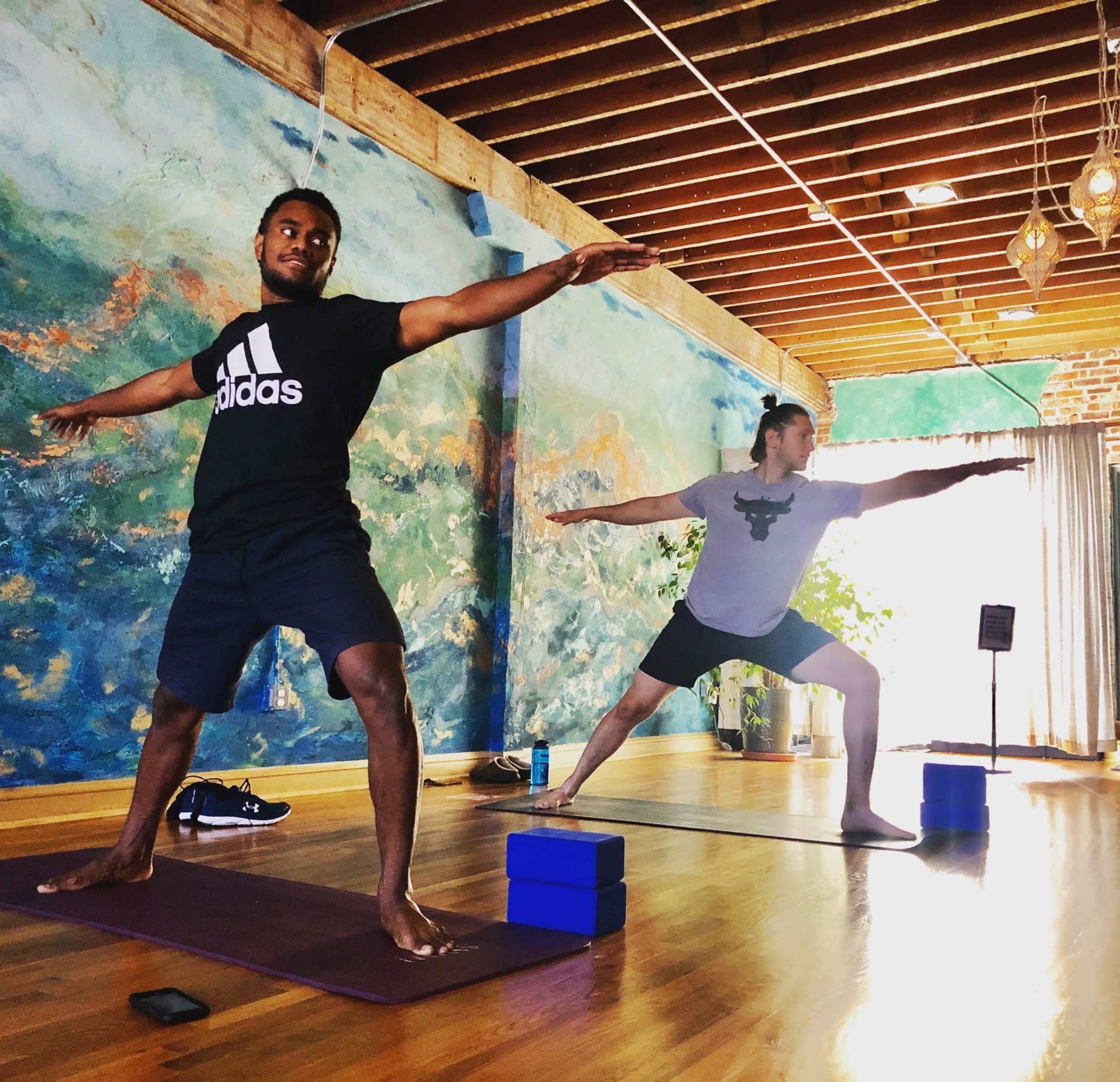 Two men perform yoga poses in a studio with a colorful mural on the wall.