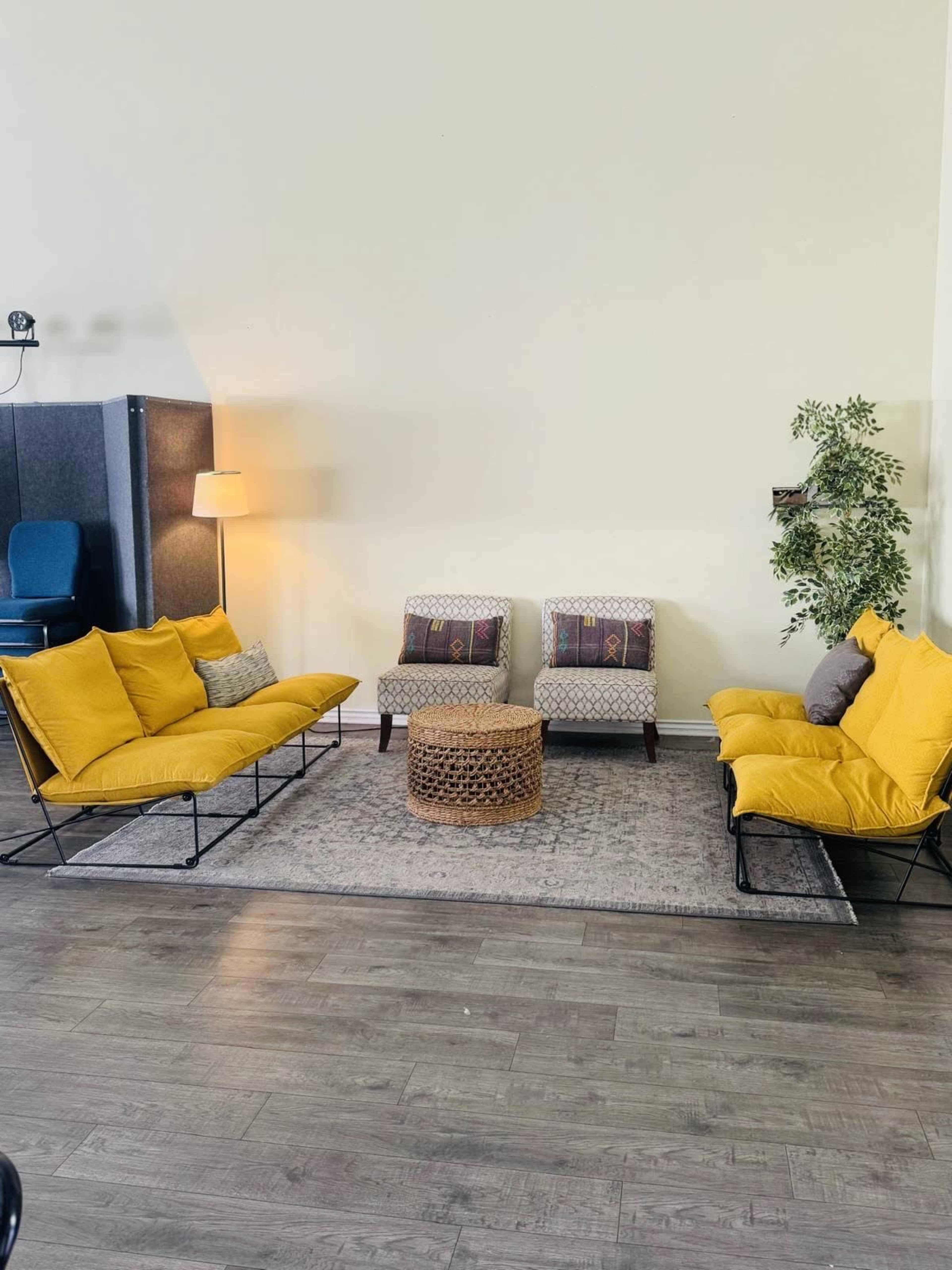 The image shows a bright, modern living area with two yellow sofas, a round wicker coffee table, and patterned accent chairs arranged on a decorative rug.