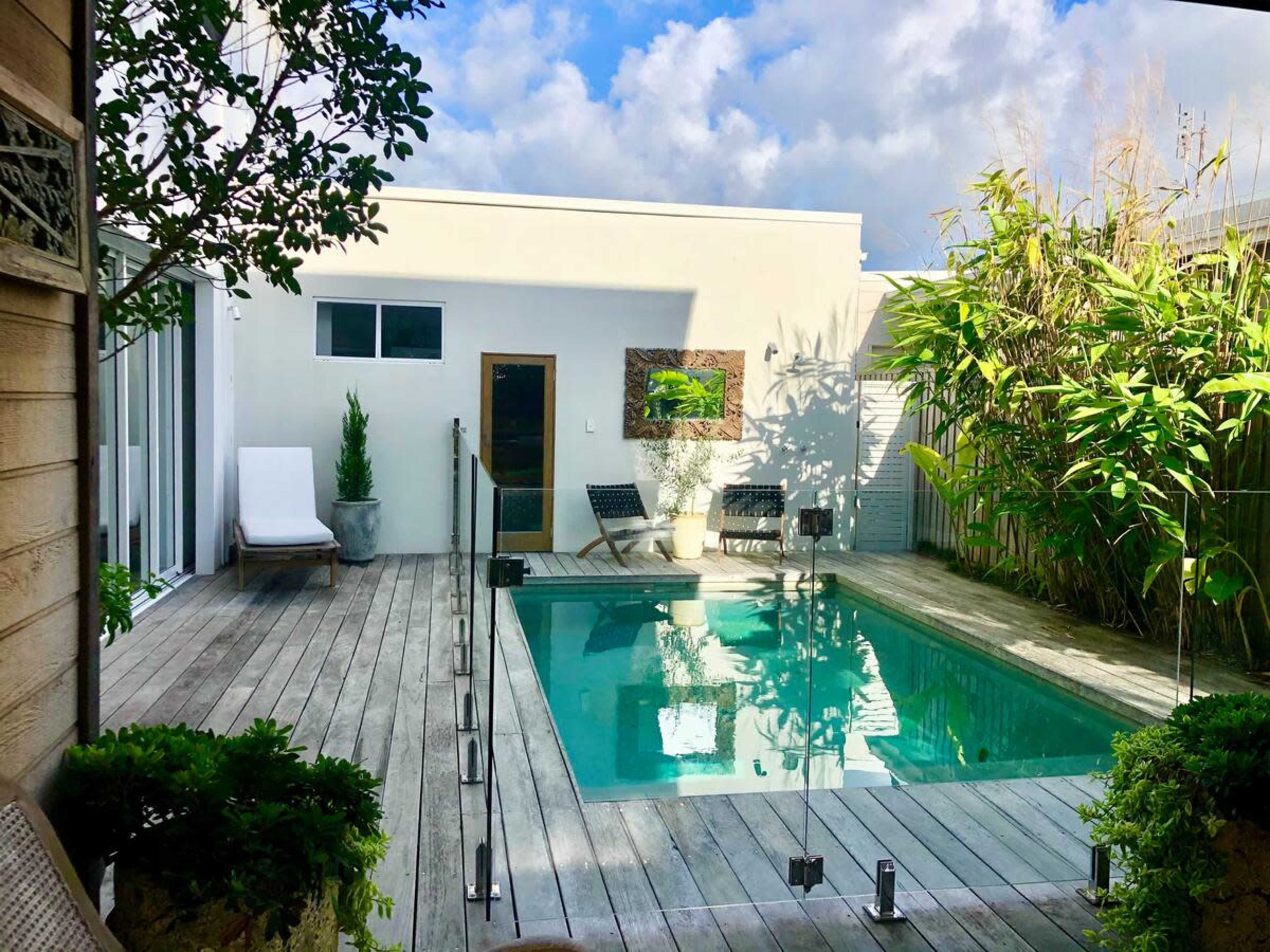 A rectangular swimming pool is surrounded by wooden decking and lush greenery in a modern backyard setting.
