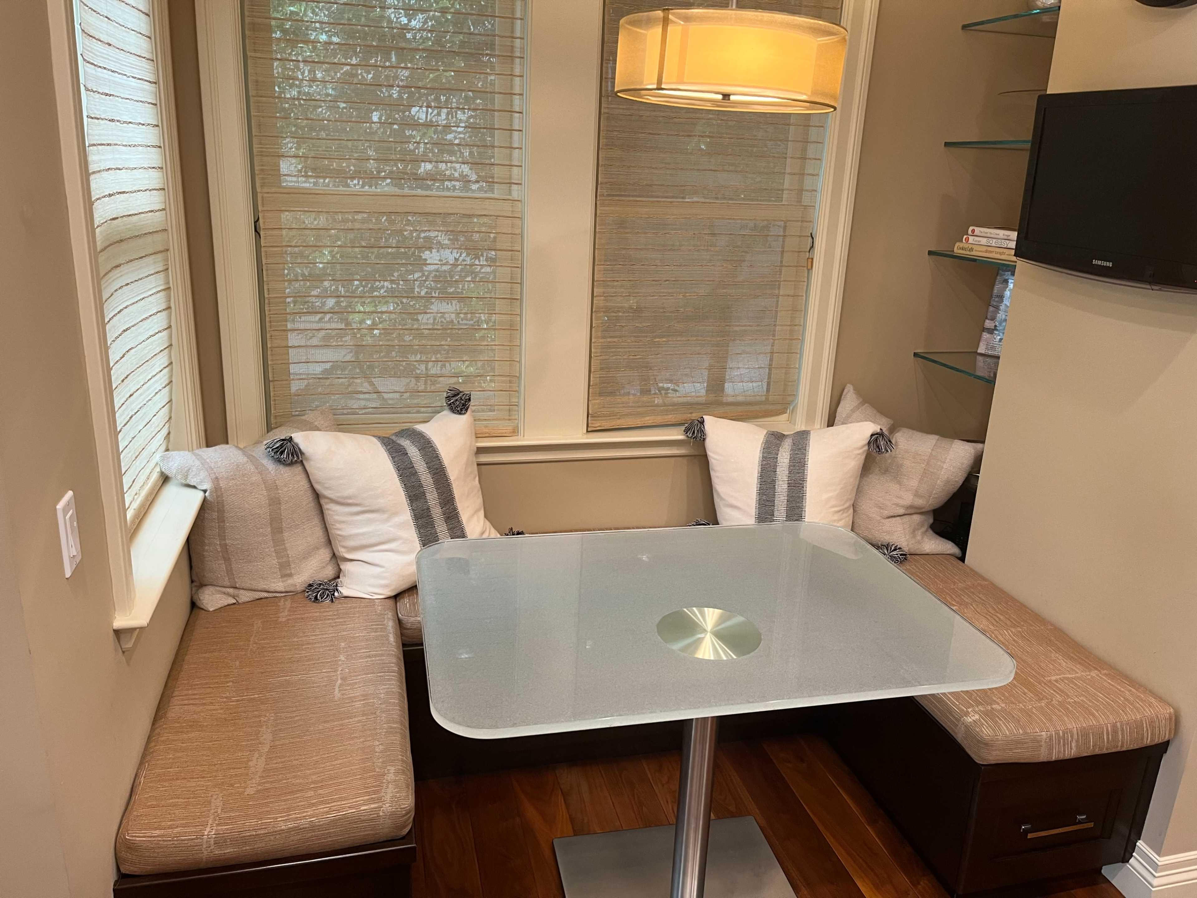 The image shows a small dining nook with a glass table and cushioned seating arranged in a corner, surrounded by windows with blinds.