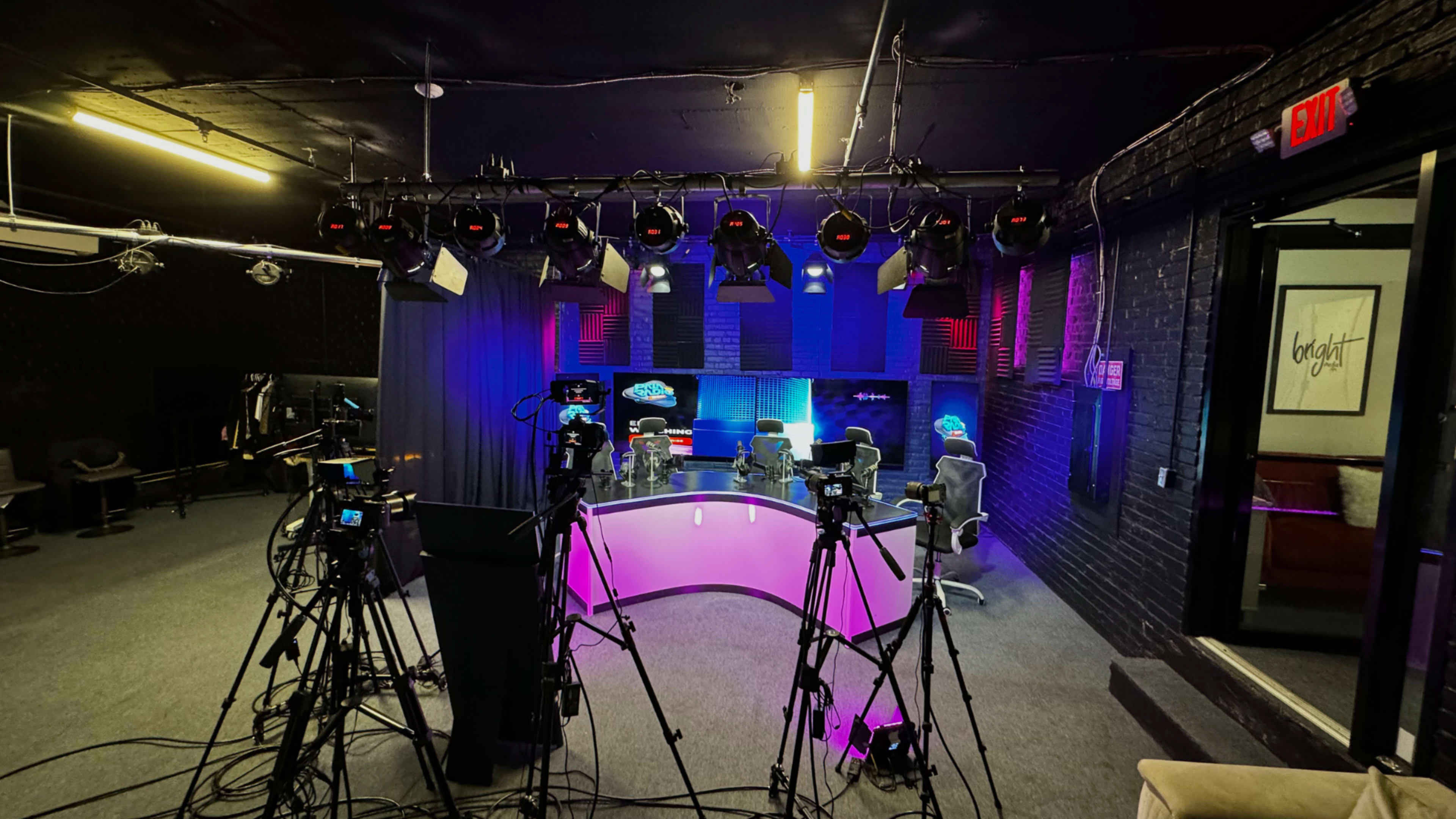 A television or streaming studio setup with multiple cameras, a curved table illuminated by pink lighting, and chairs arranged in a production area featuring colorful backdrop lighting.
