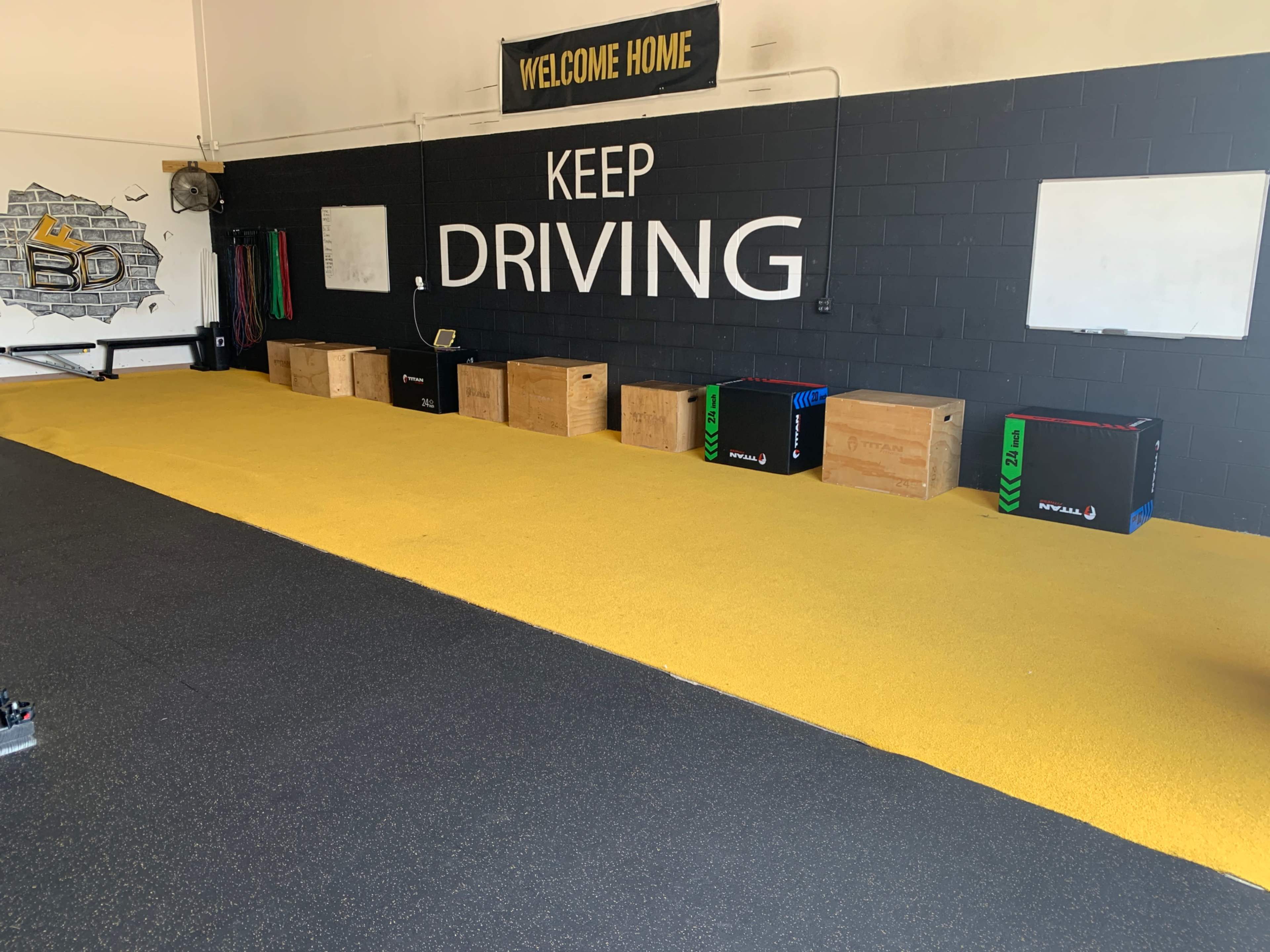 A gym interior with yellow flooring, wooden boxes lined against a black wall that has the phrase "KEEP DRIVING" prominently displayed.