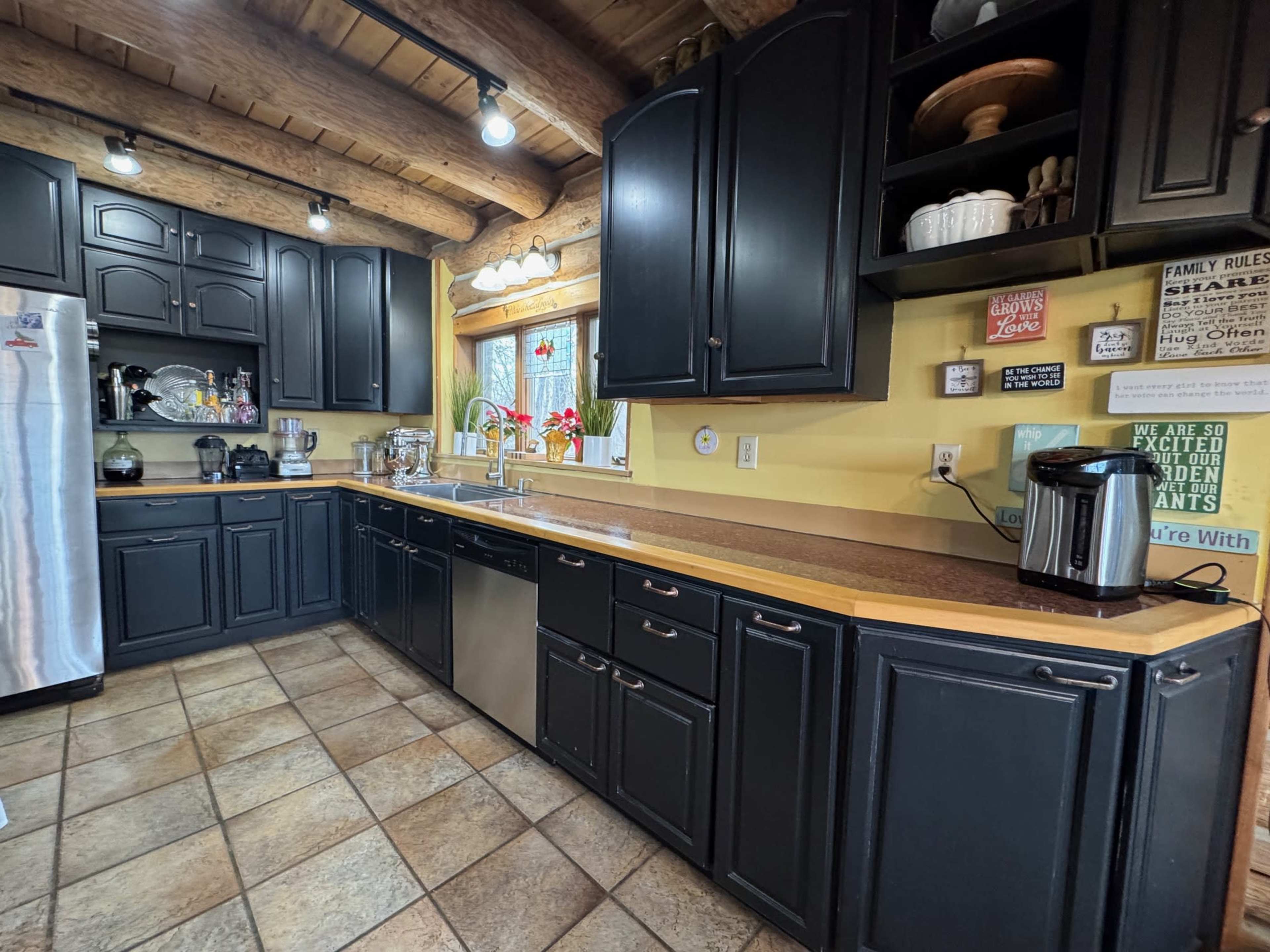 The kitchen features dark cabinetry, a stainless steel refrigerator, and a tiled floor, with natural wood beams overhead.