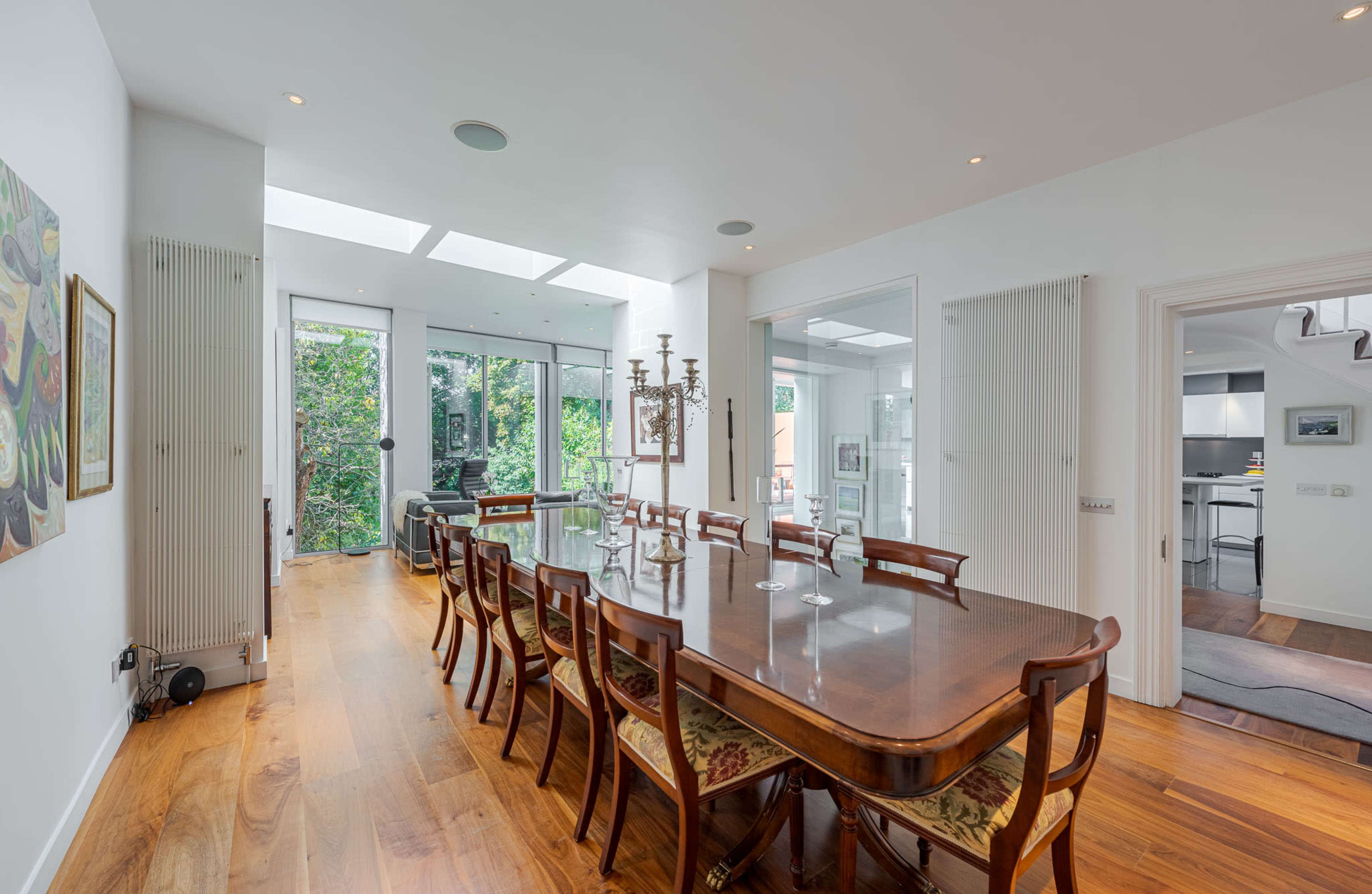 A spacious dining room features a long wooden table surrounded by chairs, with large windows that provide natural light and a view of greenery outside.