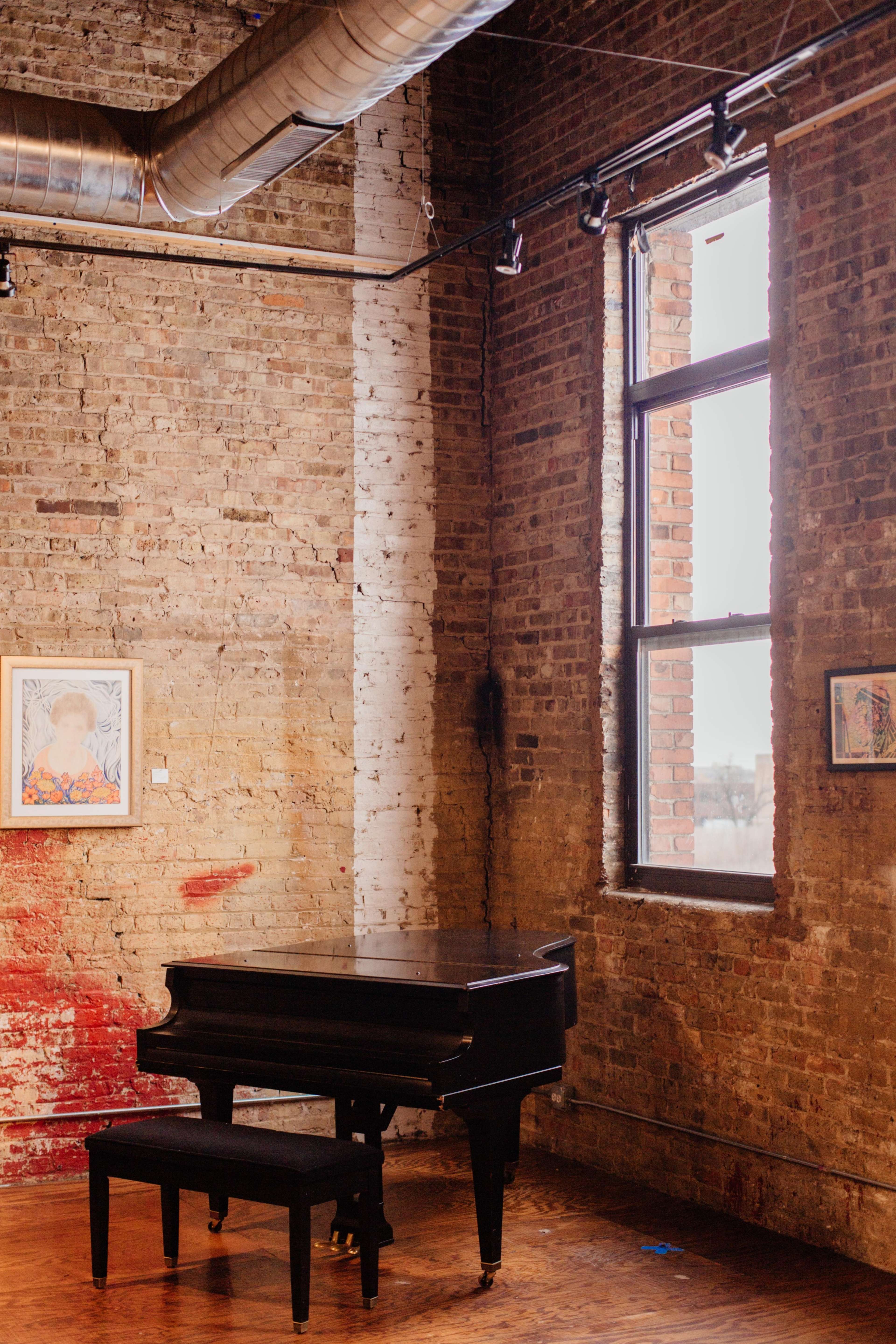 A black grand piano sits next to a window in a room with exposed brick walls and framed artwork.